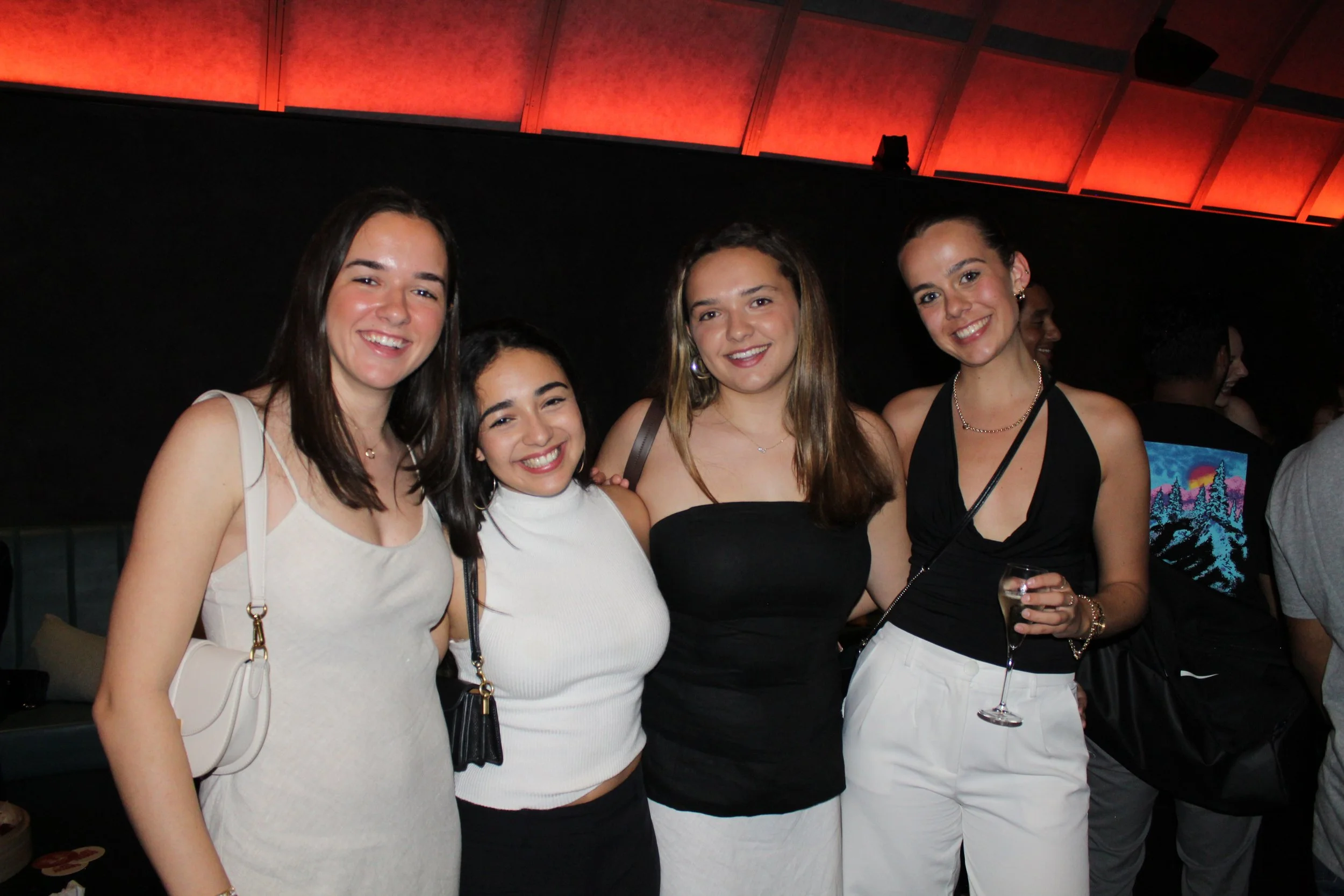 Four women smiling at a party, posing close together, with a dark background and red ceiling lights, one holding a glass of champagne.