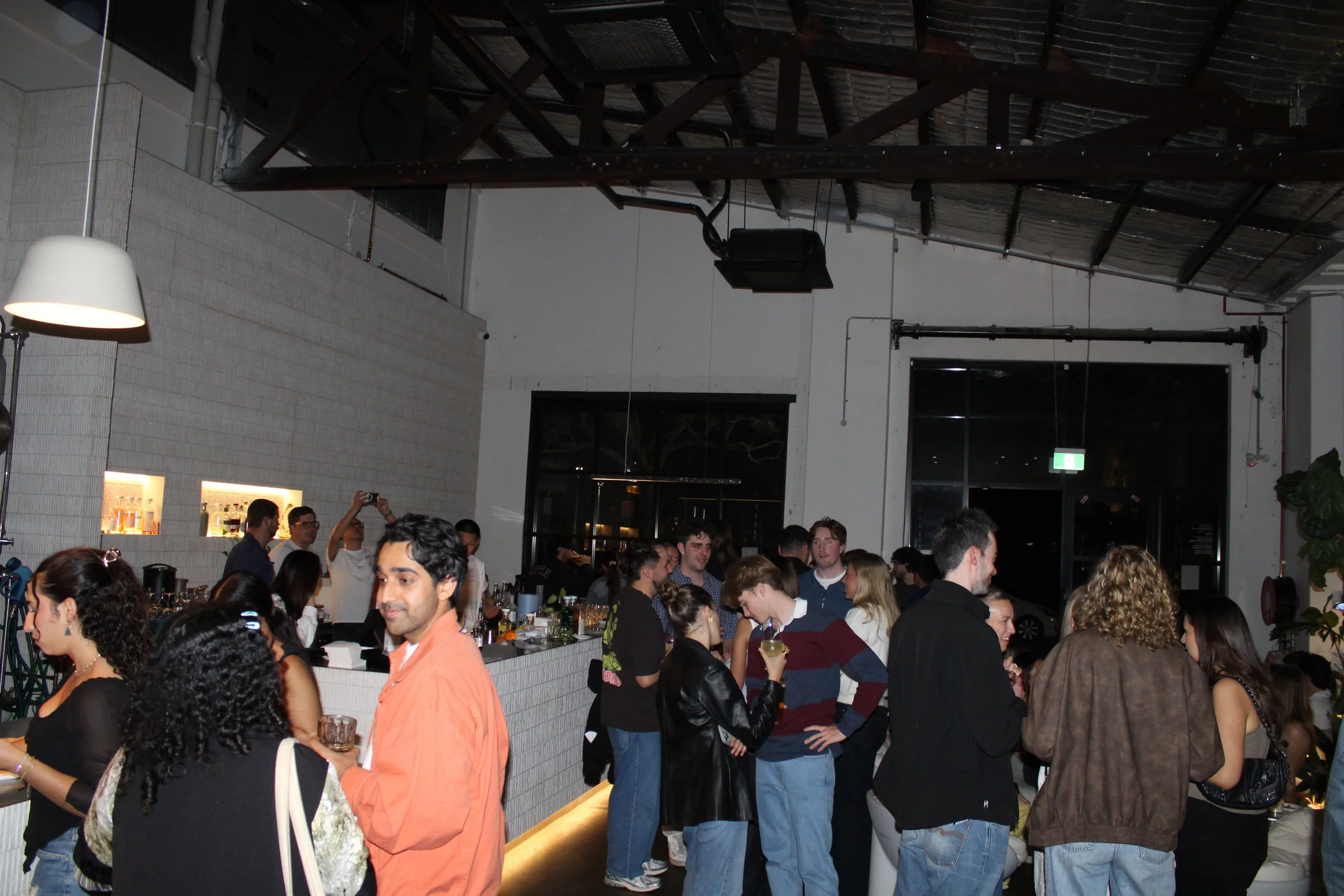 People socializing at a bar or nightclub with a modern interior, high ceiling, and dim lighting.