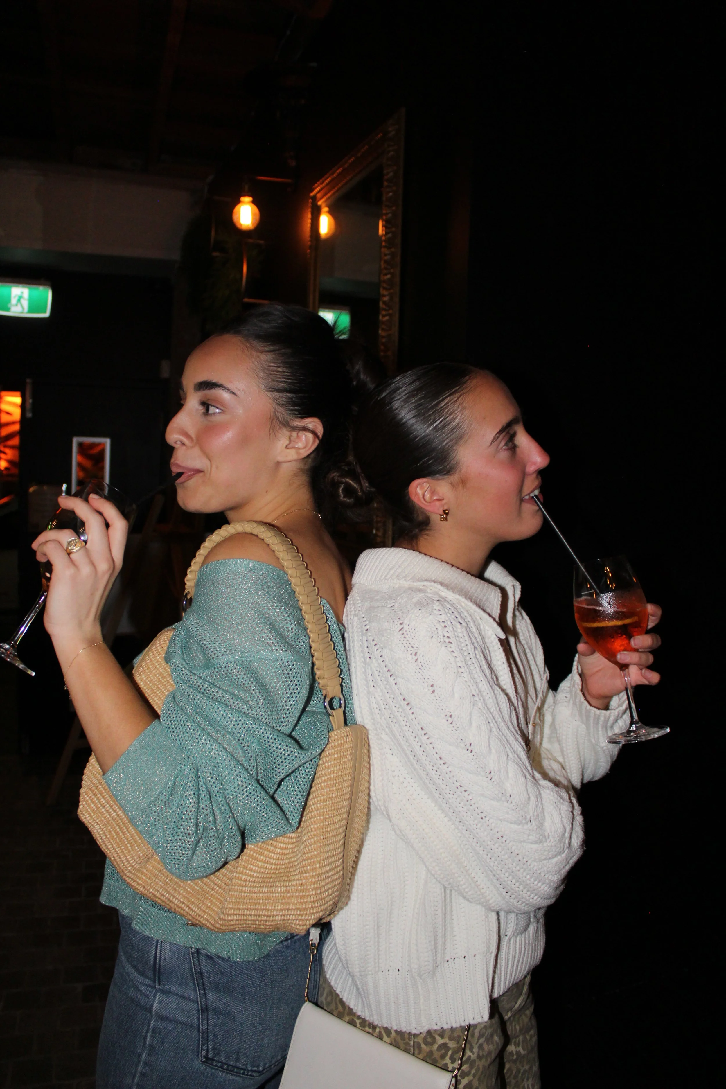 Two women standing back-to-back in a dimly lit bar or restaurant, each sipping a drink with a straw, smiling, and facing opposite directions.