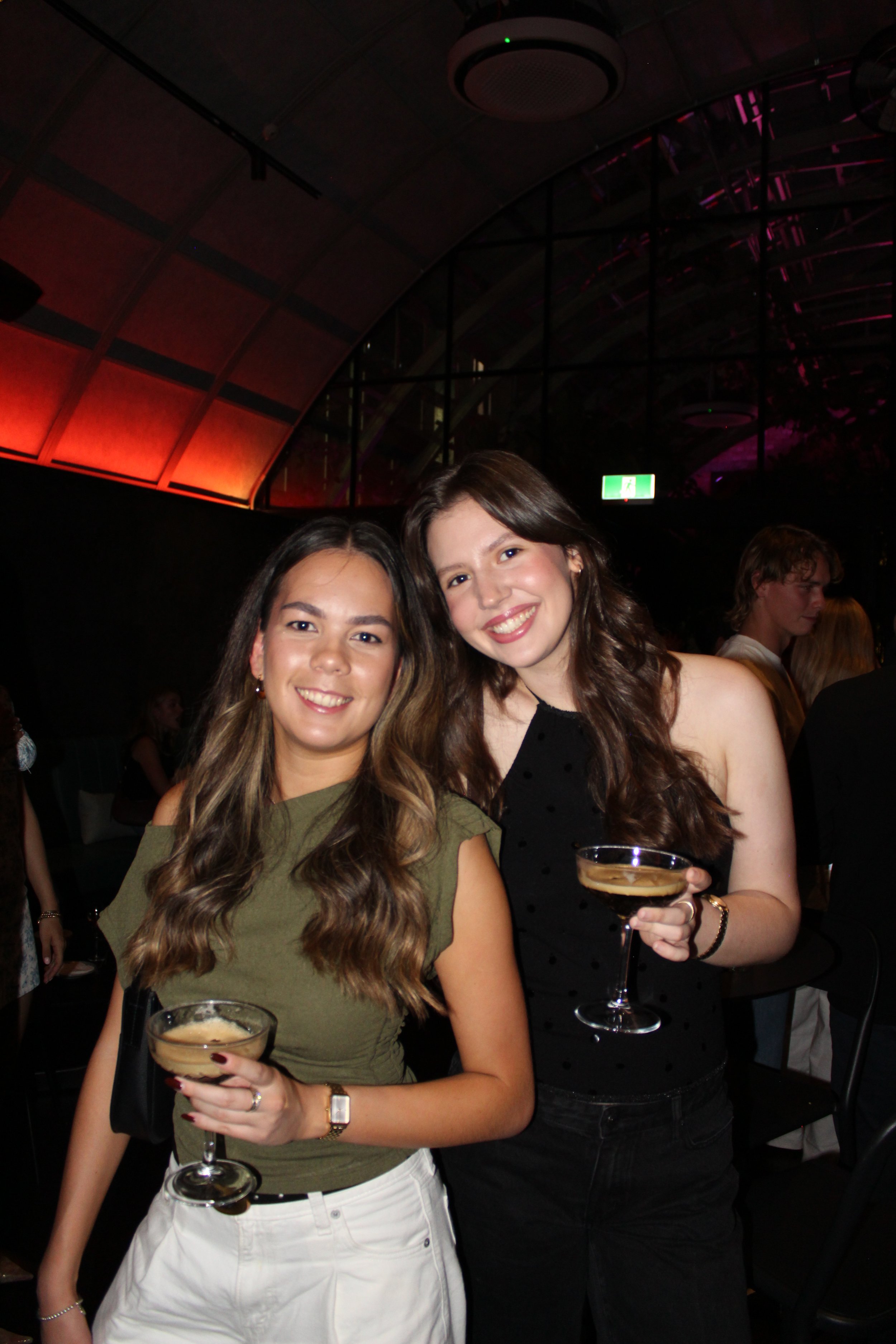 Two young women smiling and holding cocktail glasses at a party or social event.