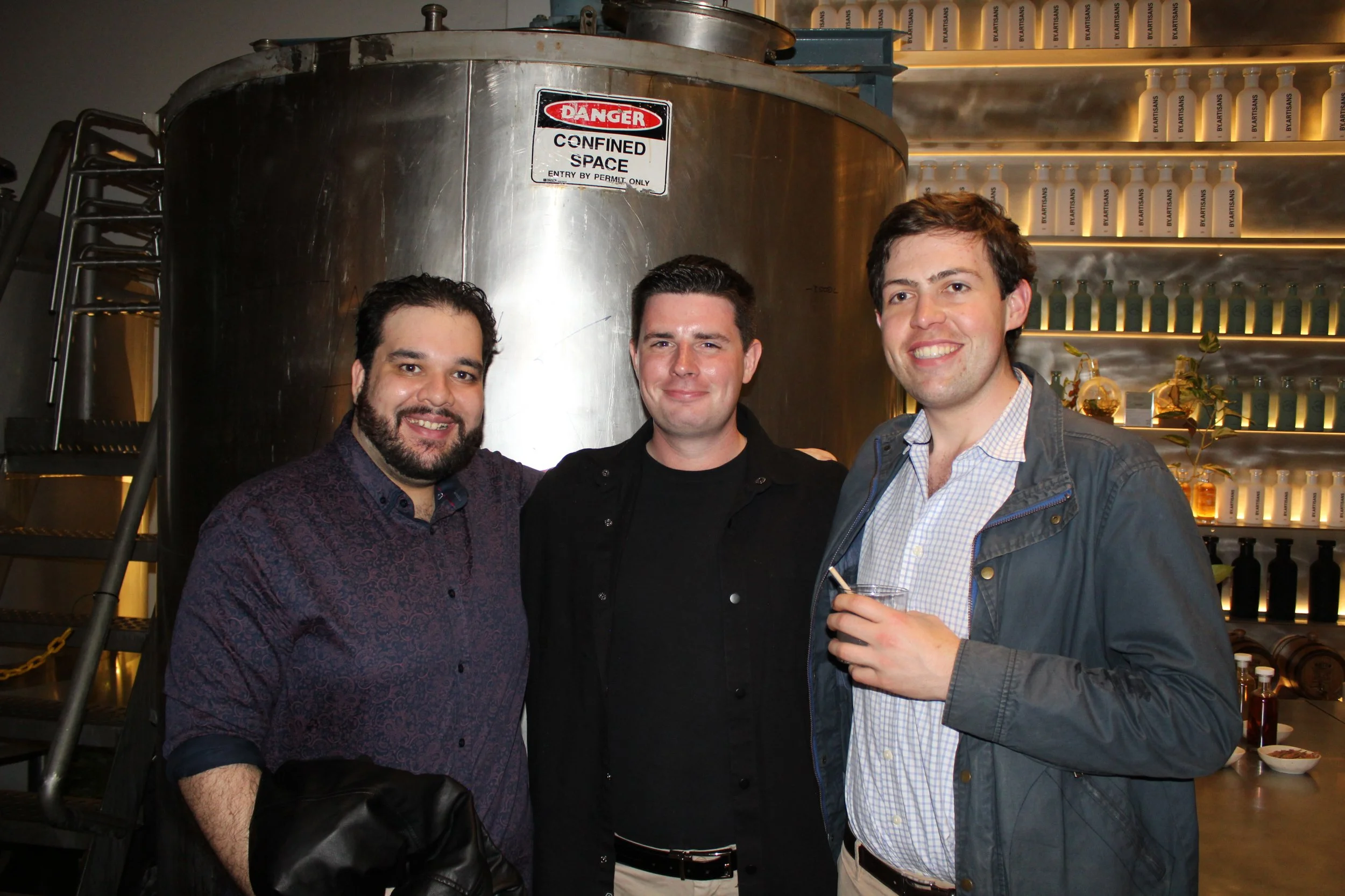Three men standing together in a brewery or bar, smiling at the camera, with metal brewing equipment and shelves of bottles in the background.