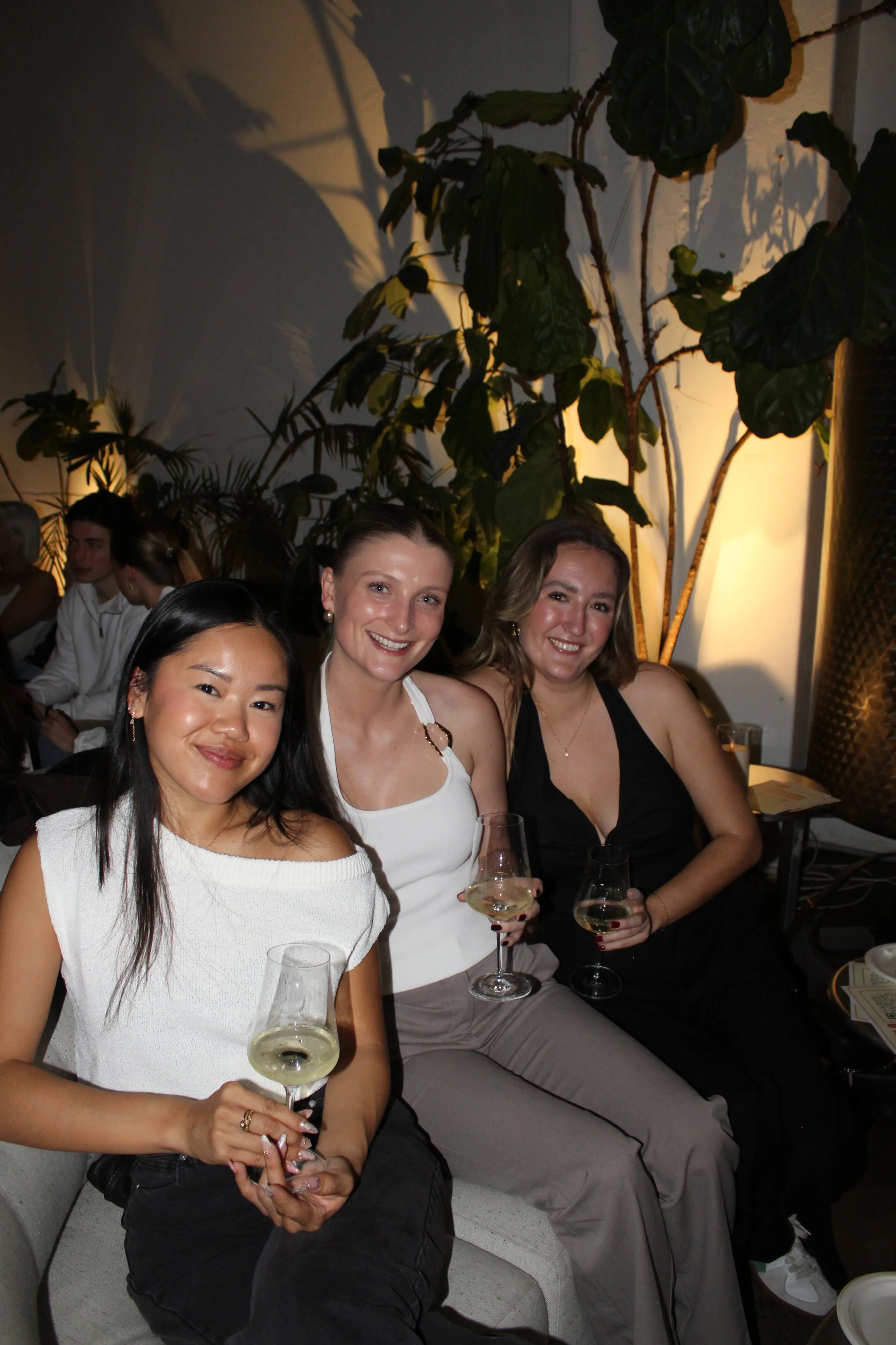 Three women sitting and smiling, holding glasses of white wine, in a dimly lit indoor setting with large green plants in the background.