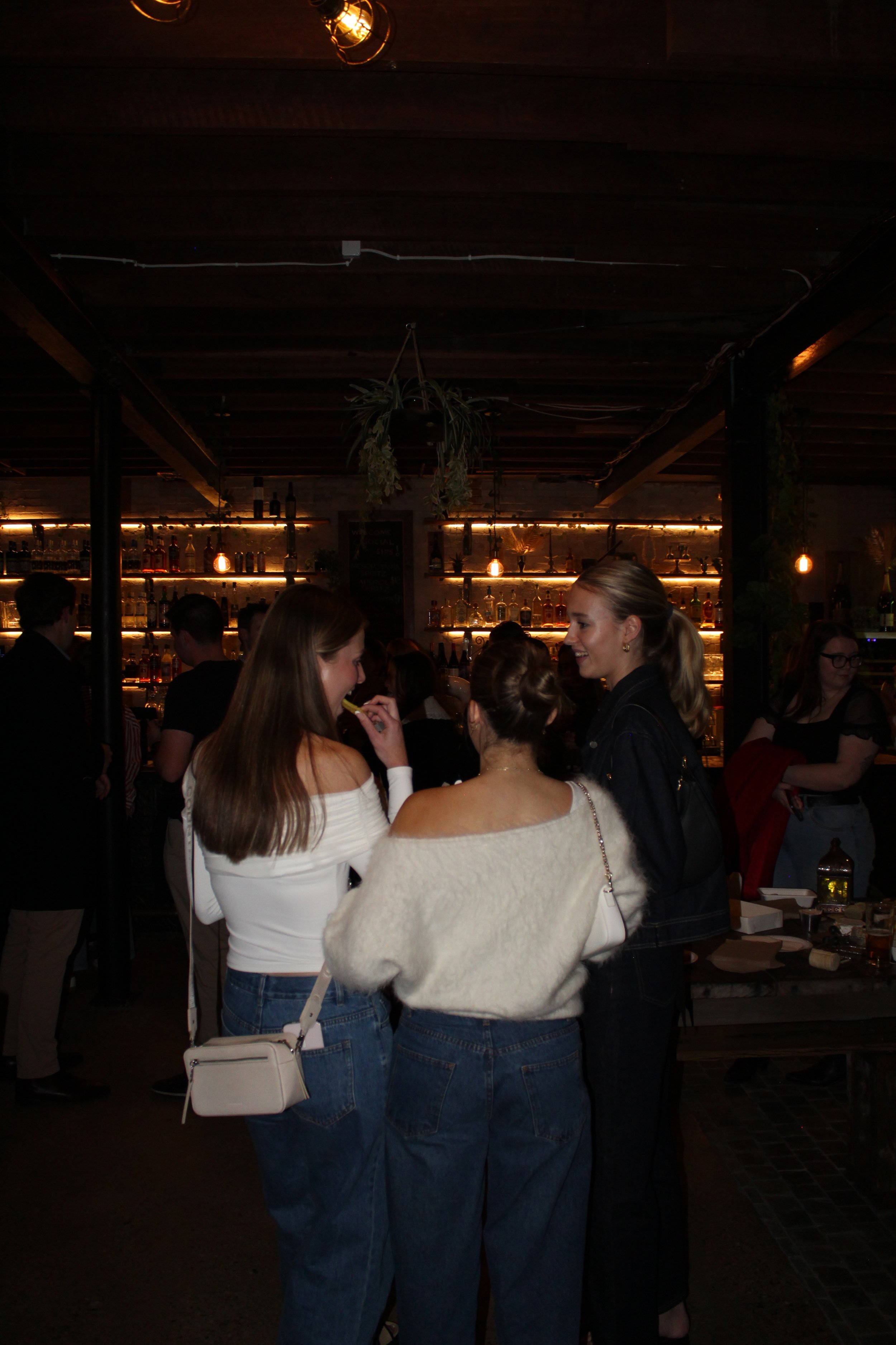 Three women stand and chat in a dimly lit bar or restaurant with bottles on shelves in the background.