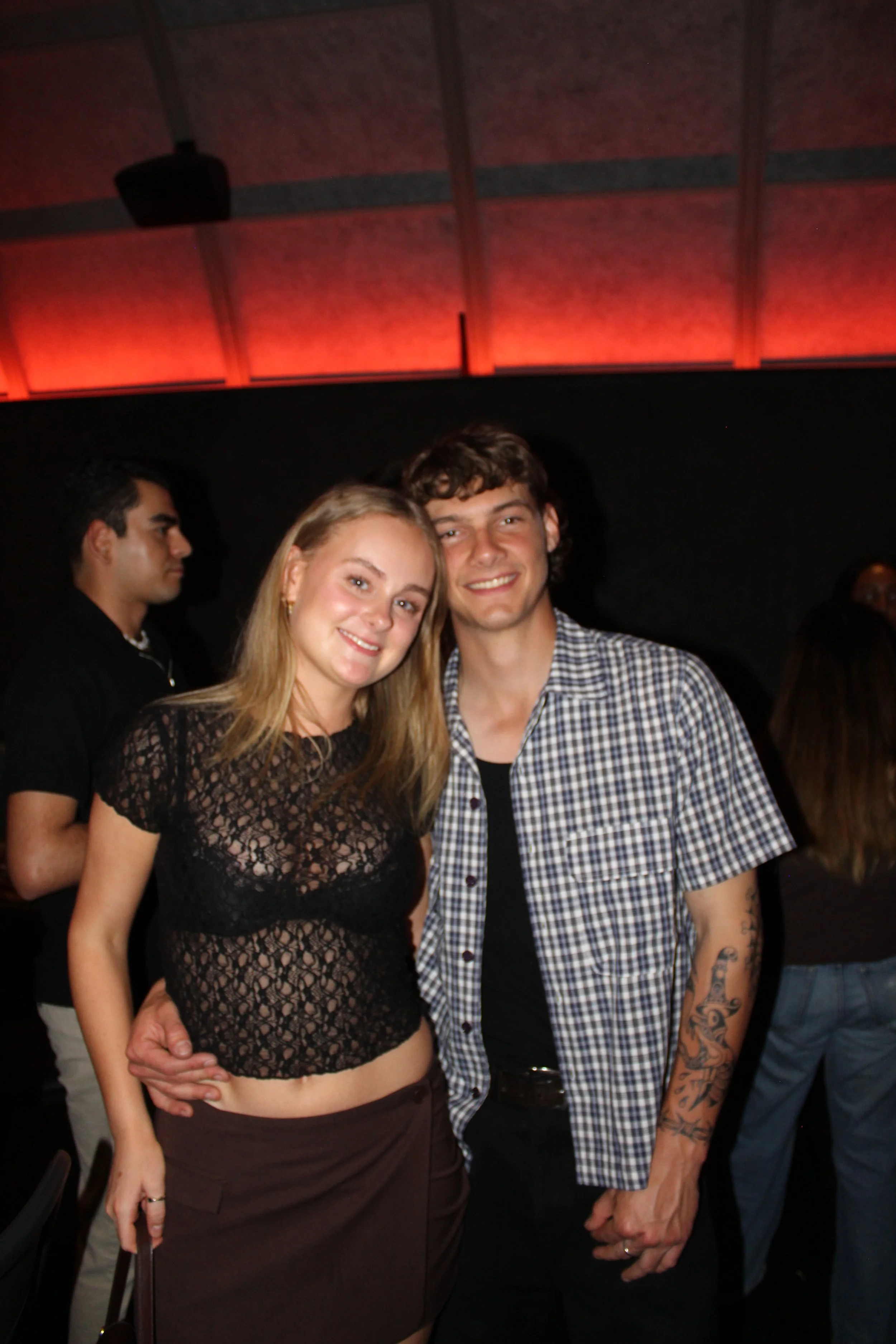 A young woman and young man smiling, standing close together indoors at a social event. The woman wears a black lace crop top, and the man wears a short-sleeved plaid shirt with tattoos on his arm.