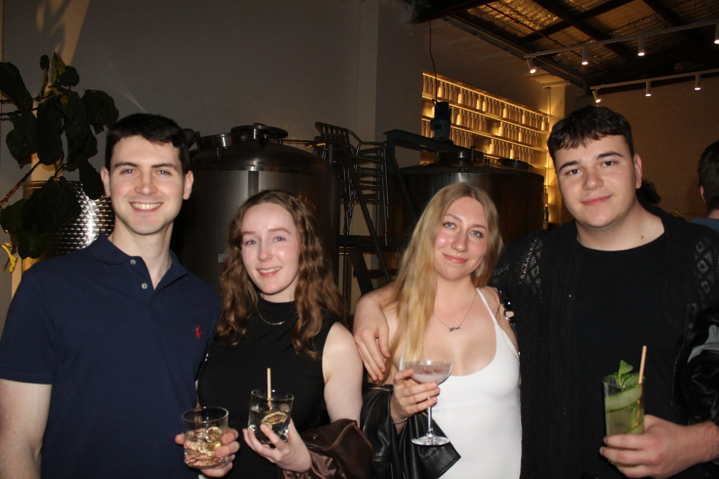 Four young adults, two men and two women, smiling and holding drinks at a social gathering in a modern indoor venue.