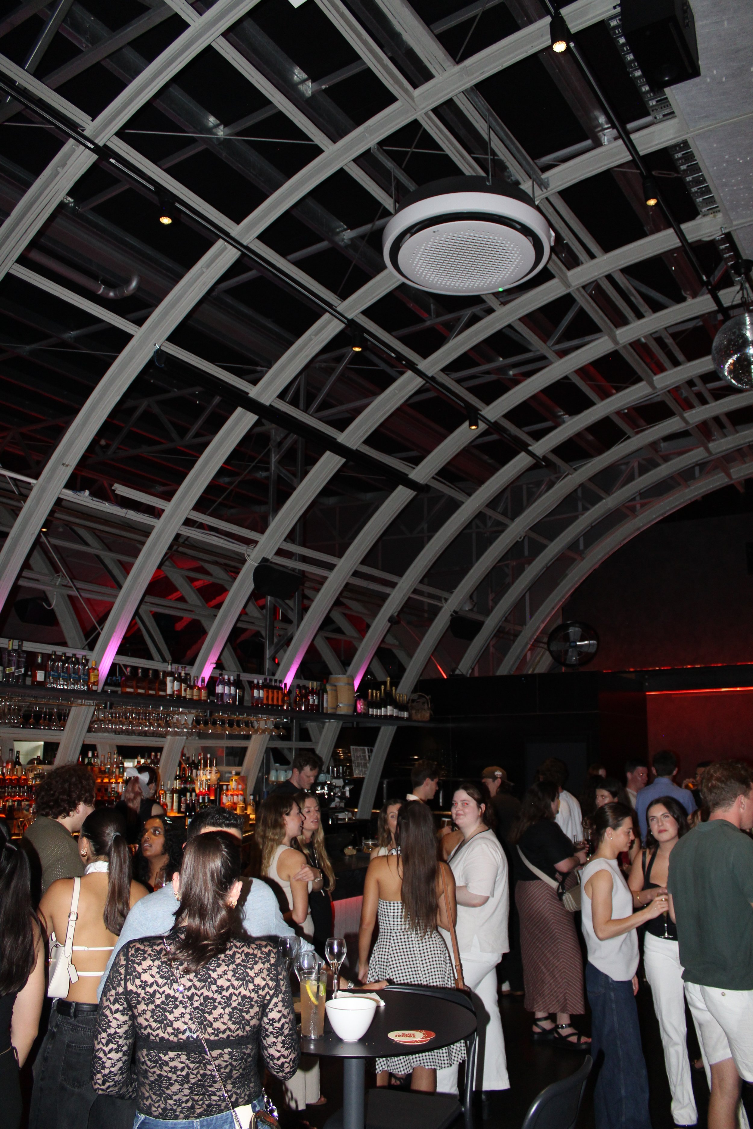 A crowded indoor bar or nightclub with people socializing, some holding drinks, under an arched ceiling with exposed beams and modern lighting fixtures.