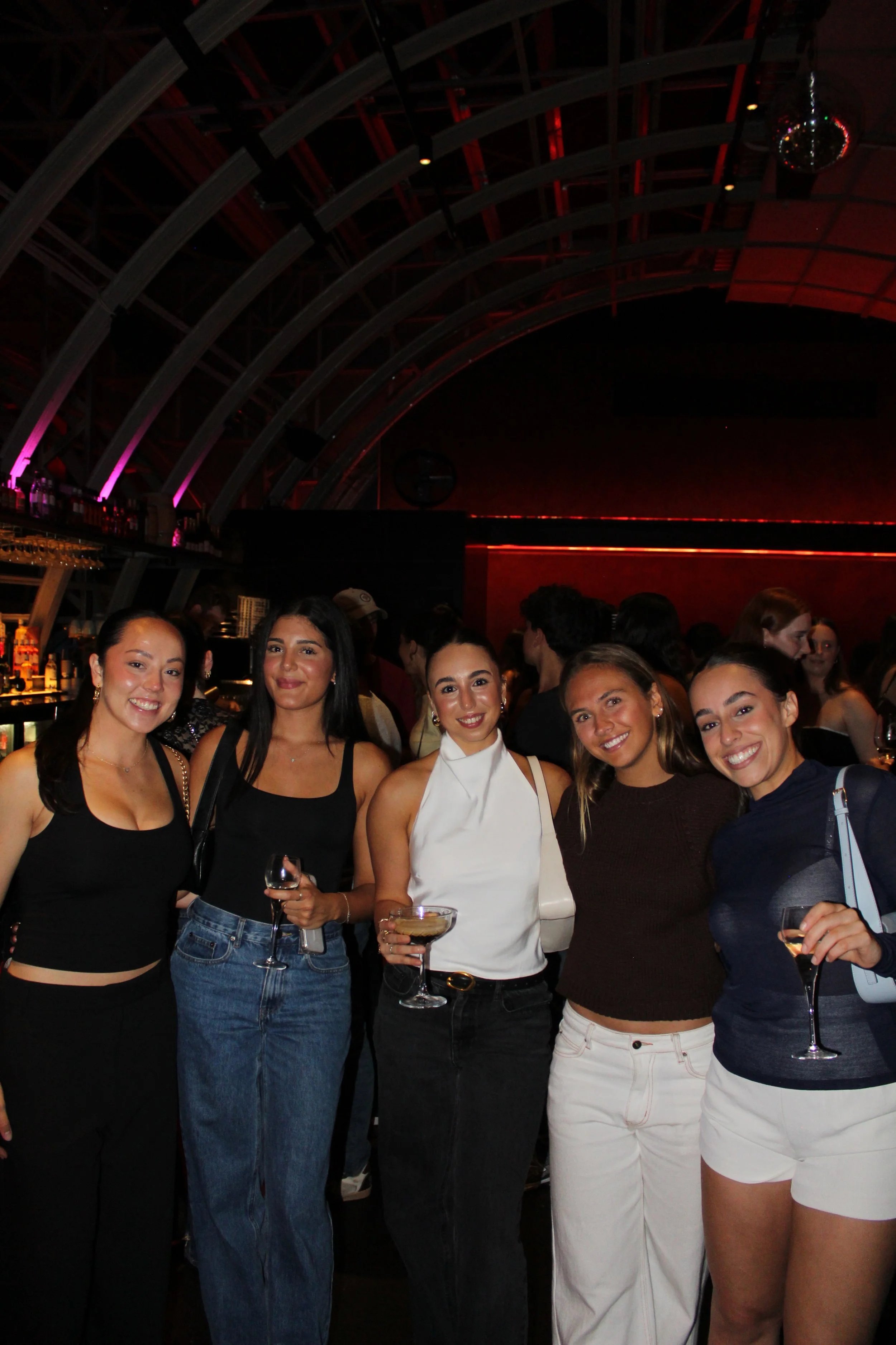 Group of five women smiling and posing with drinks at a lively bar or club with dim lighting and a decorated ceiling.
