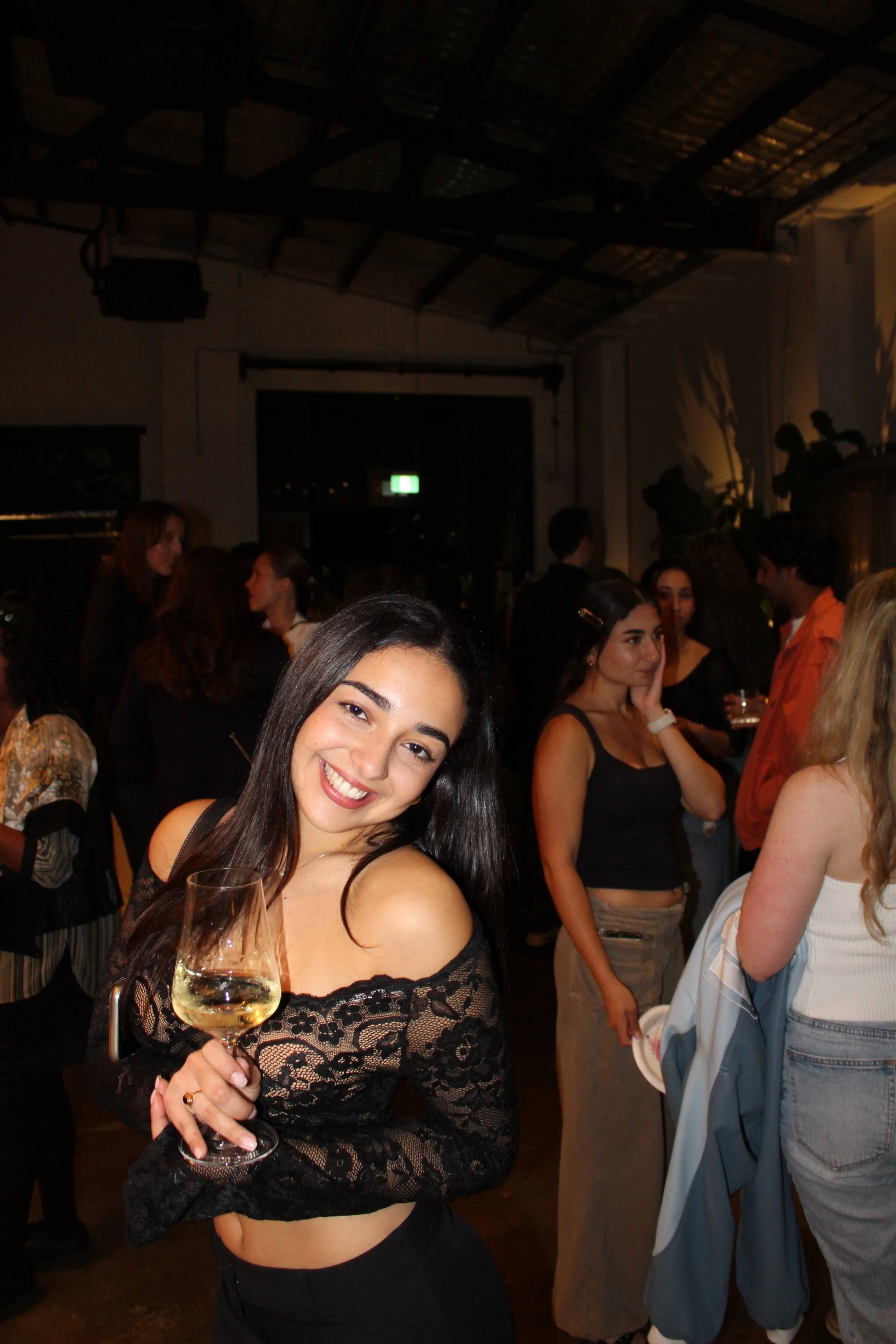 A young woman with long dark hair smiling and holding a glass of white wine at a social gathering or party.