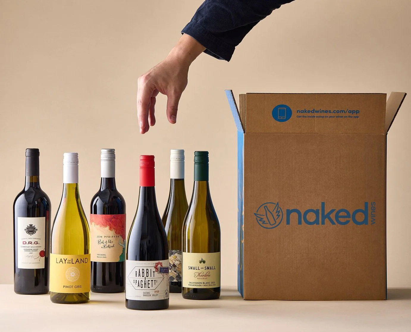 Array of six wine bottles with various labels and colors being picked up from a table by a hand, next to a cardboard box with the 'nakedwines' logo and website on it, against a plain background.
