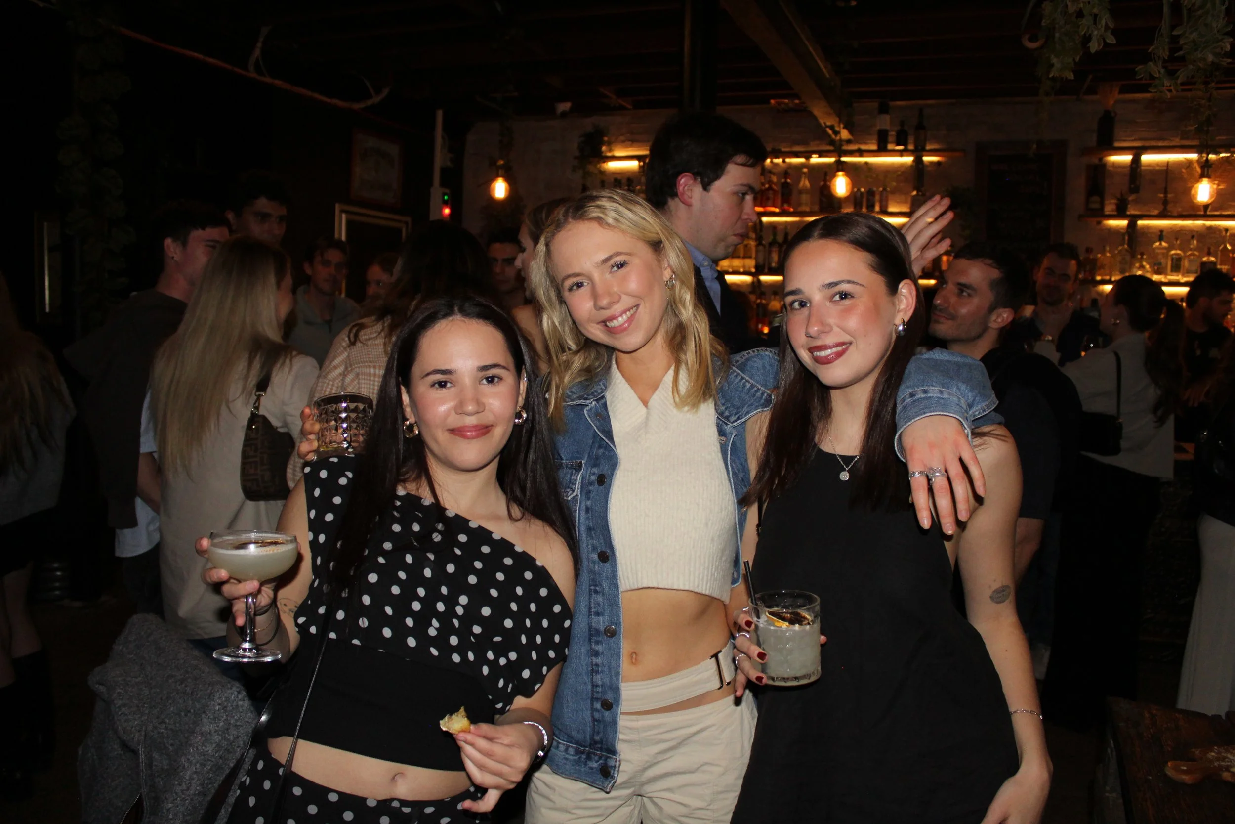 Three young women posing for a photo at a bar or nightclub, holding drinks, with a group of people in the background.