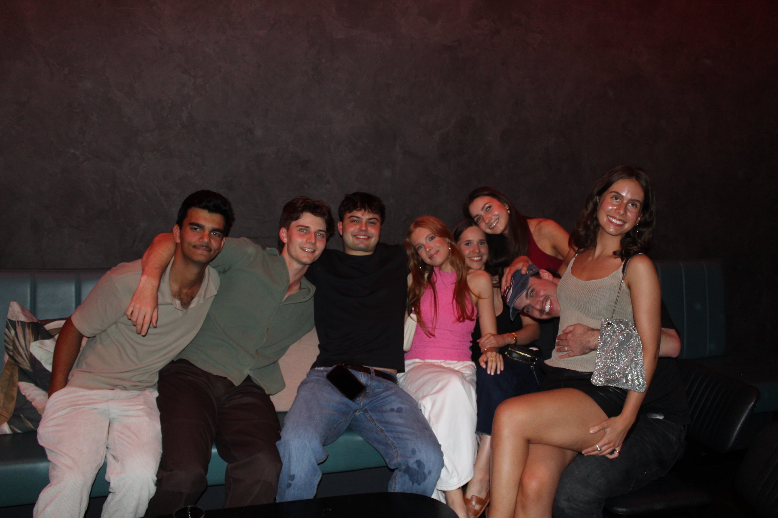 Group of eight young adults sitting closely together on a green leather sofa, smiling and enjoying each other's company in a dimly lit setting.