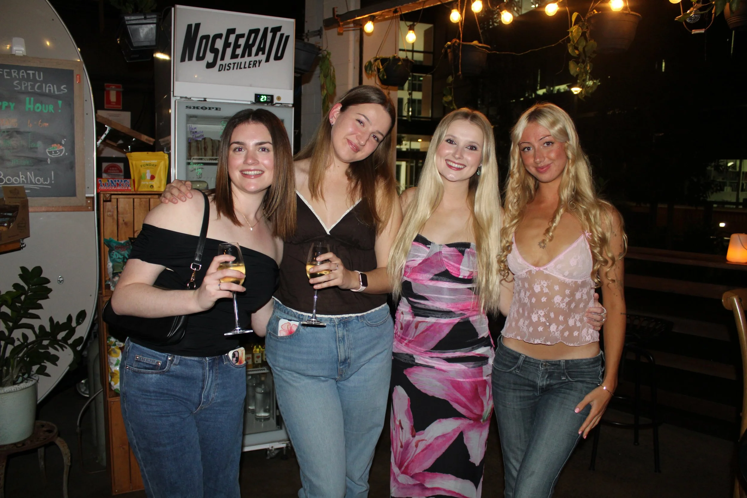 Four women standing together at a bar or restaurant, smiling and holding glasses of white wine.