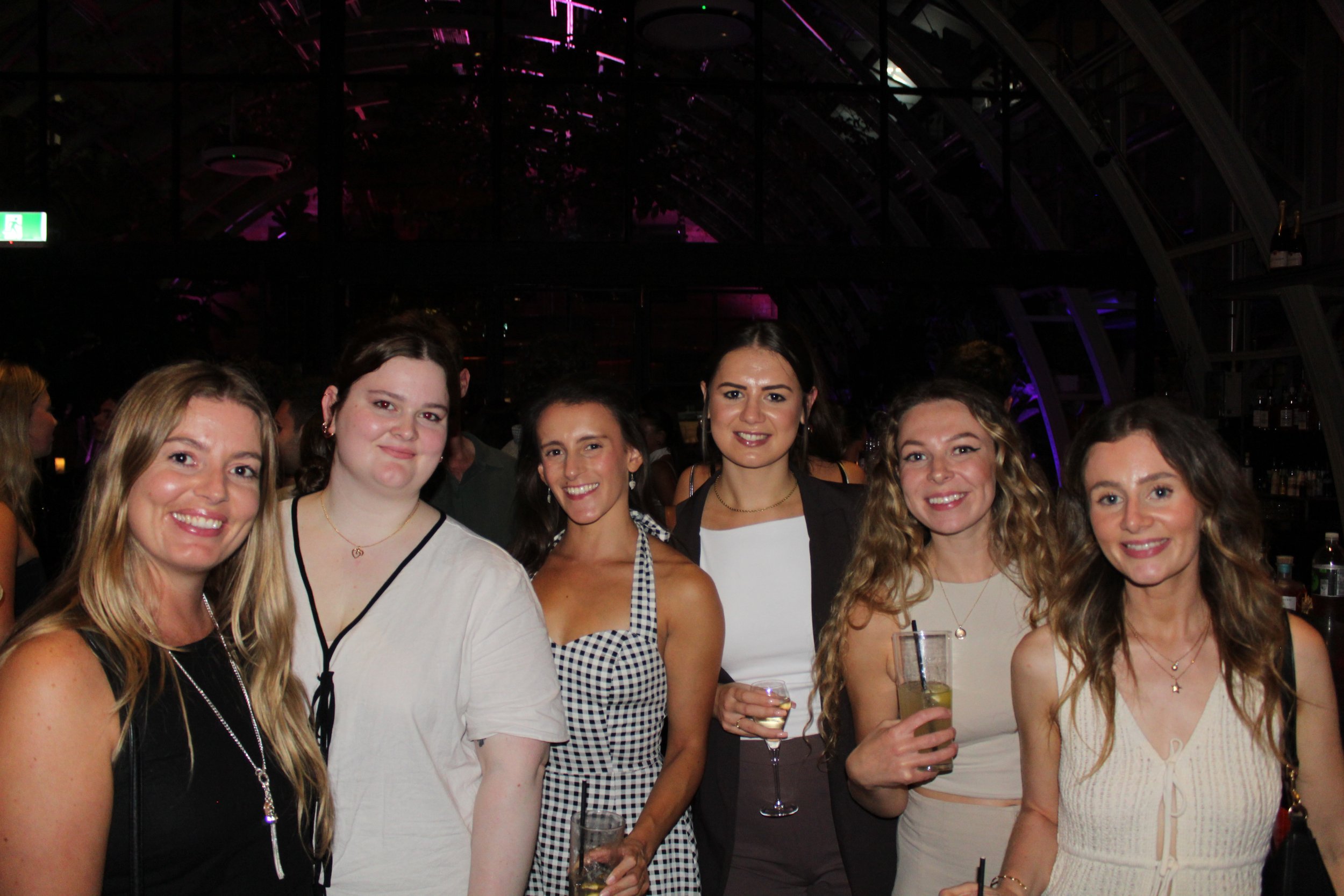 Six women at a party or social gathering, smiling and holding drinks, with a dark indoor setting and purple lighting in the background.
