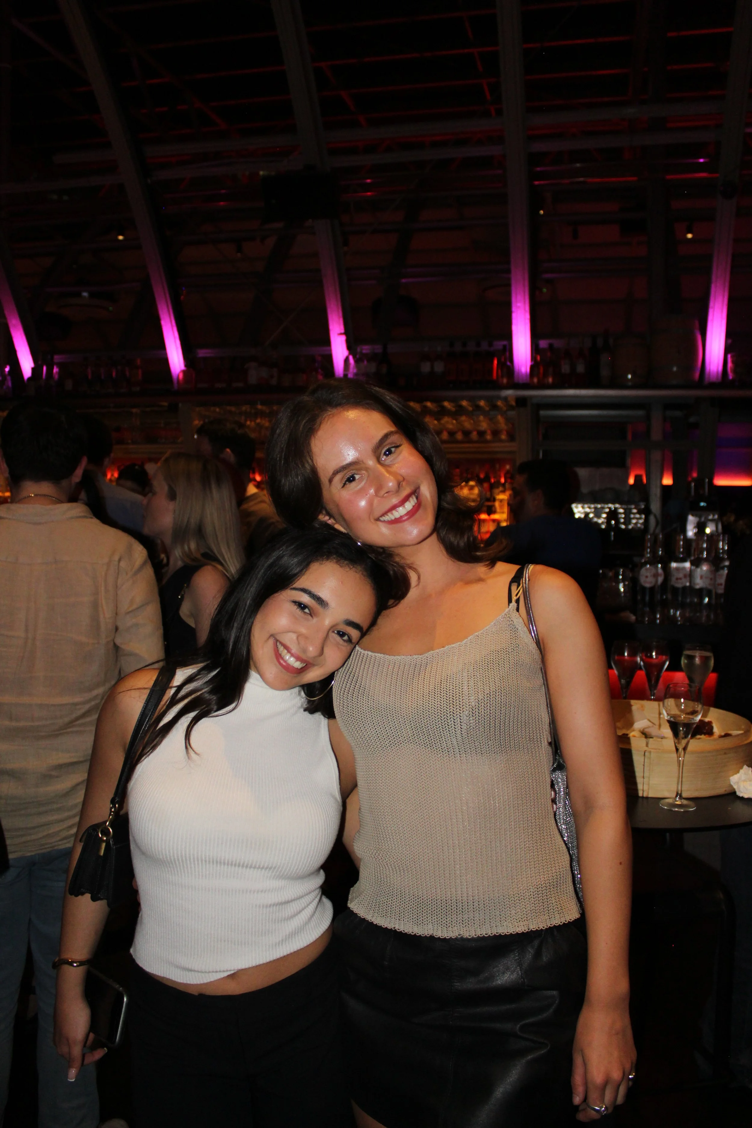 Two women smiling at a crowded bar or nightclub with purple and pink lighting in the background.