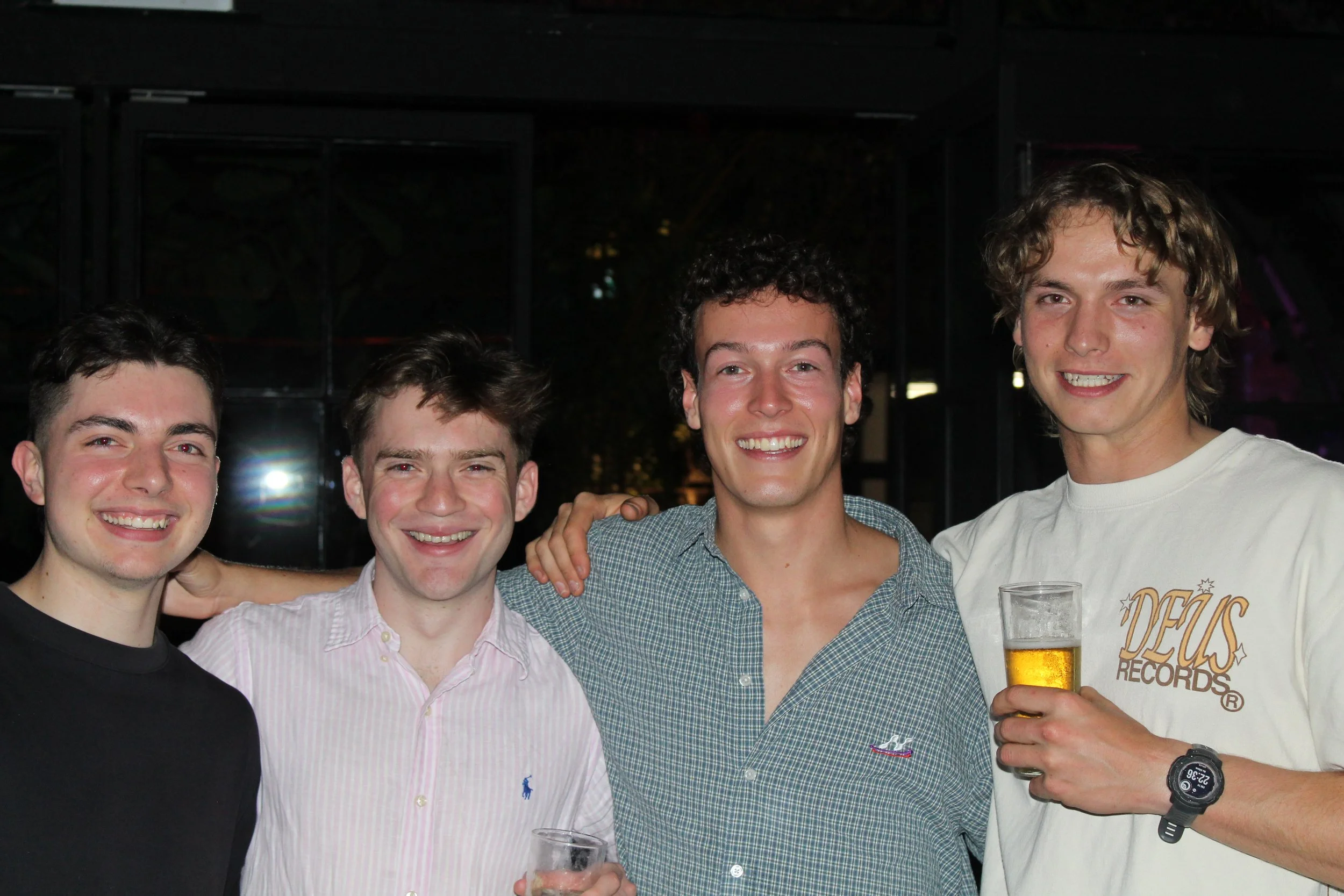 Four young men smiling and posing for a photo together at a social gathering, with two holding glasses of beer.