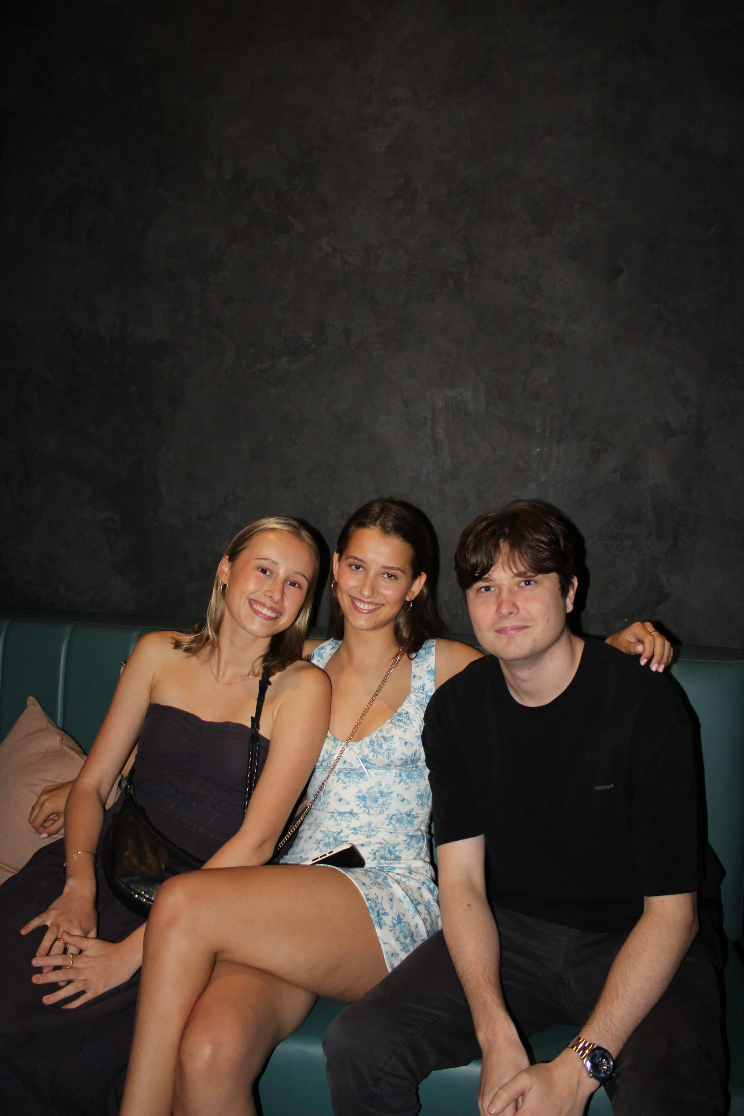 Three friends sitting on a teal booth in a dark indoor setting, smiling at the camera.