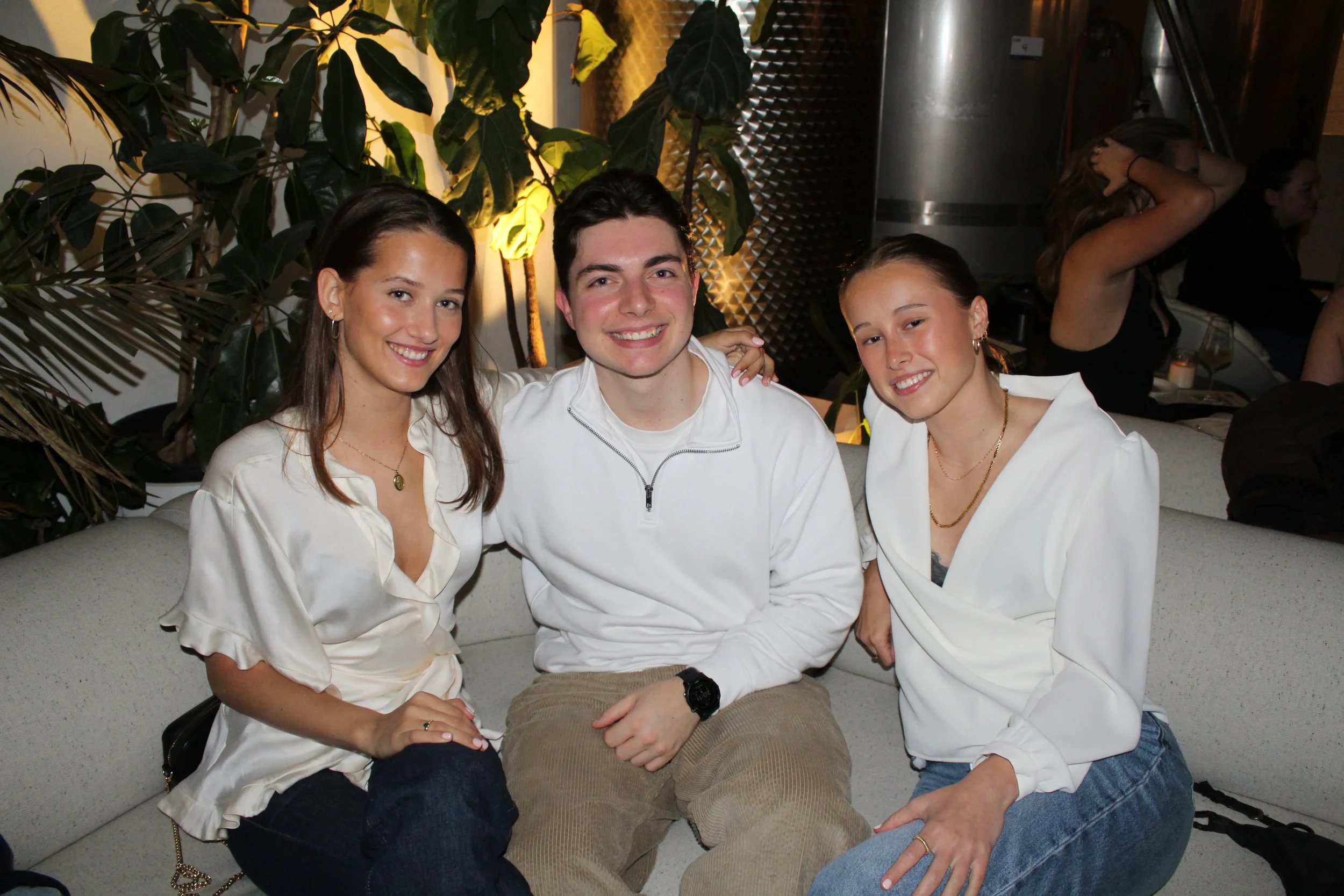 Three young adults, two women and one man, sitting on a light-colored couch at a social gathering or party, smiling at the camera. The women wear white blouses, and the man wears a white shirt with a zip collar. There are large indoor plants and othe