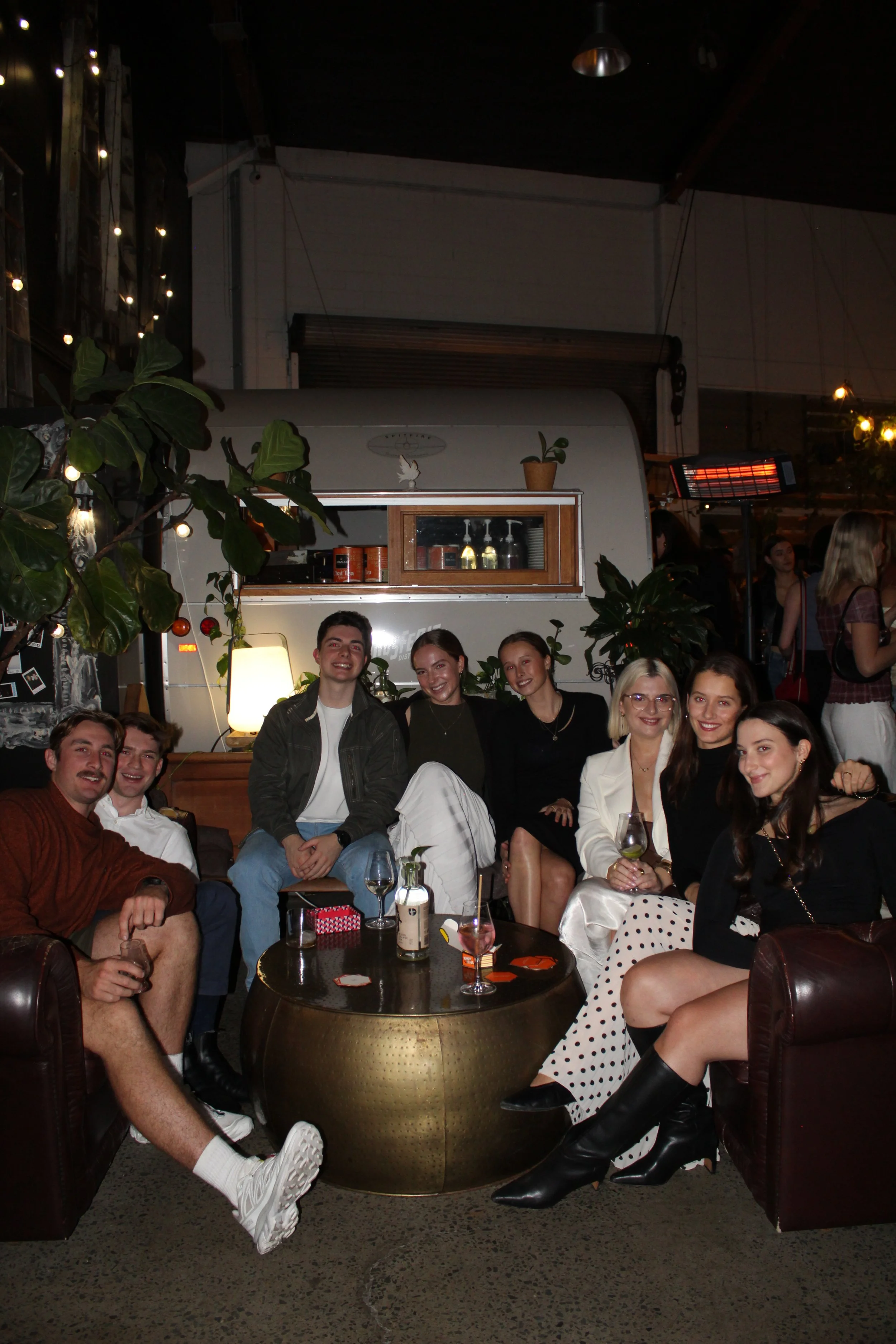Group of nine people sitting and standing around a gold coffee table in a dimly lit indoor space, with a white caravan in the background, plants, and string lights.