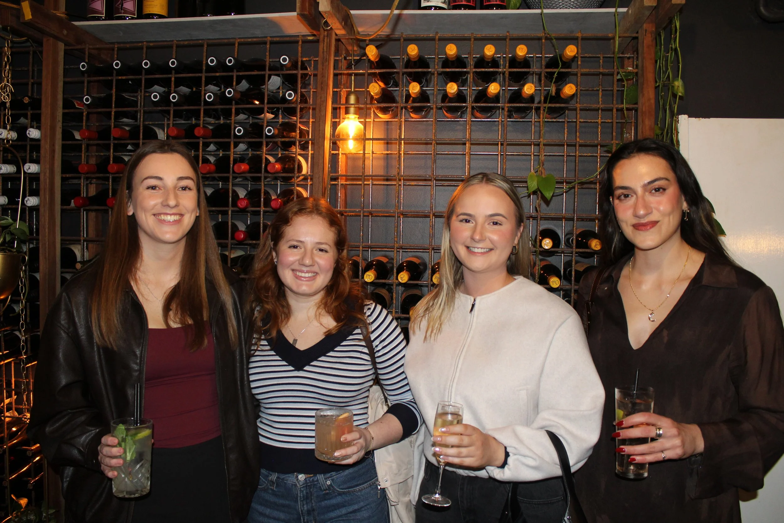 Four women smiling and holding drinks at a social gathering in a bar or restaurant, with a wine rack and plants in the background.