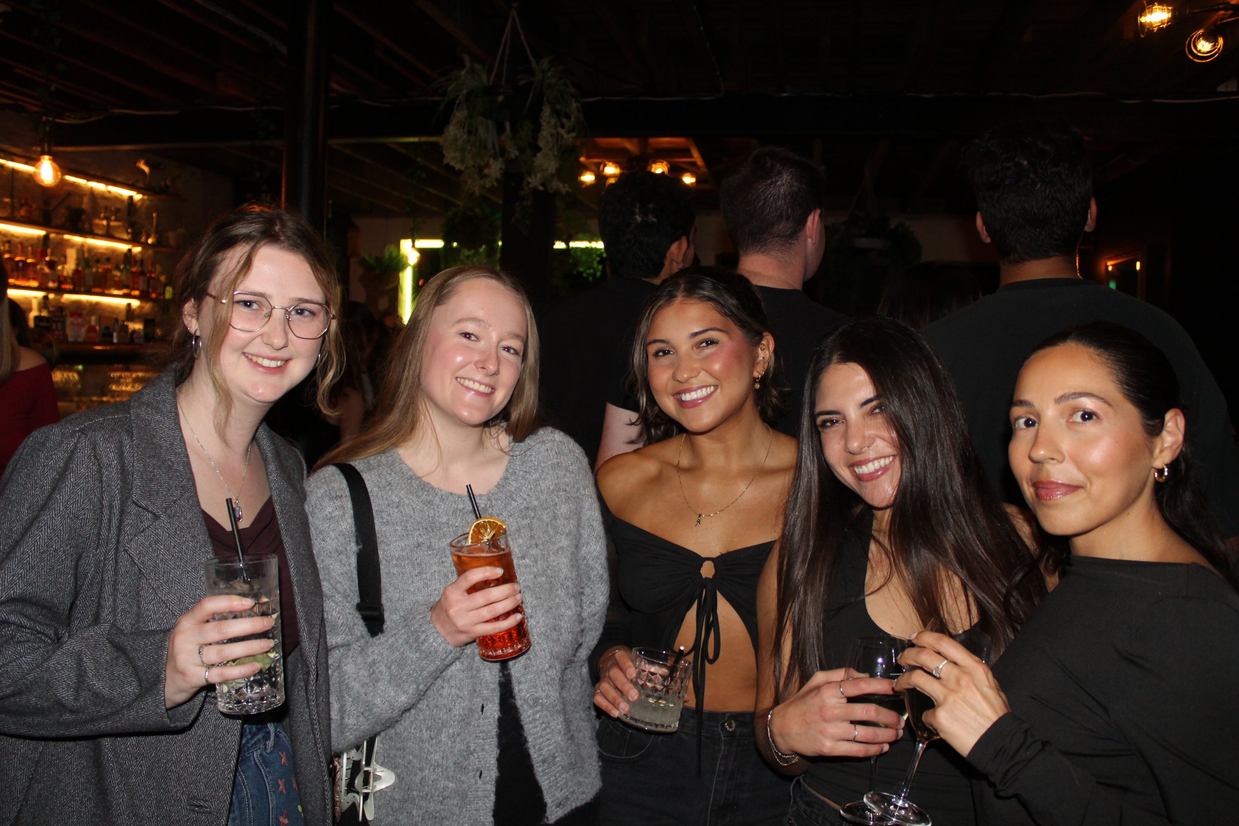 Five women in a bar holding drinks, smiling at the camera, with a dimly lit background of shelves and hanging lights.