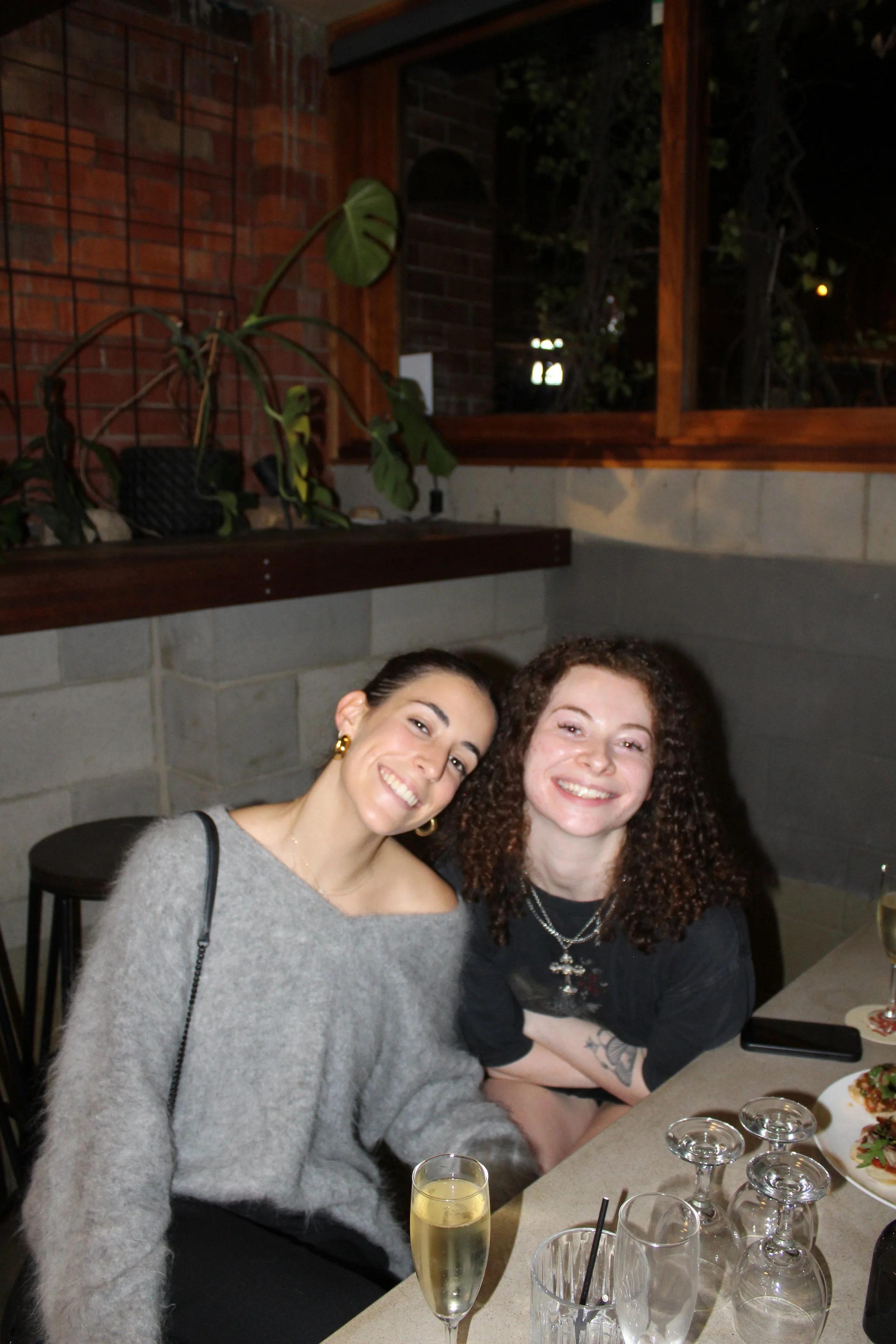 Two women smiling at a restaurant table with drinks and food, dark brick wall and plant in background.