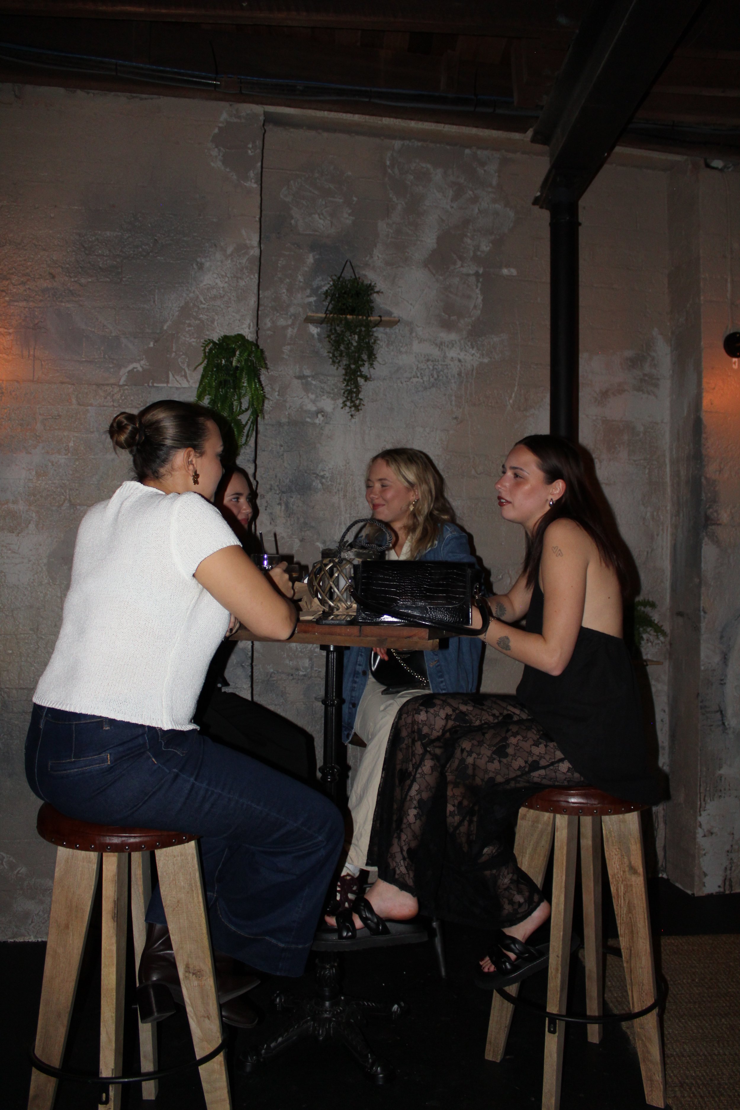 Four women sitting around a high table, chatting and drinking in a cozy, dimly lit bar or cafe with exposed brick and hanging plants.