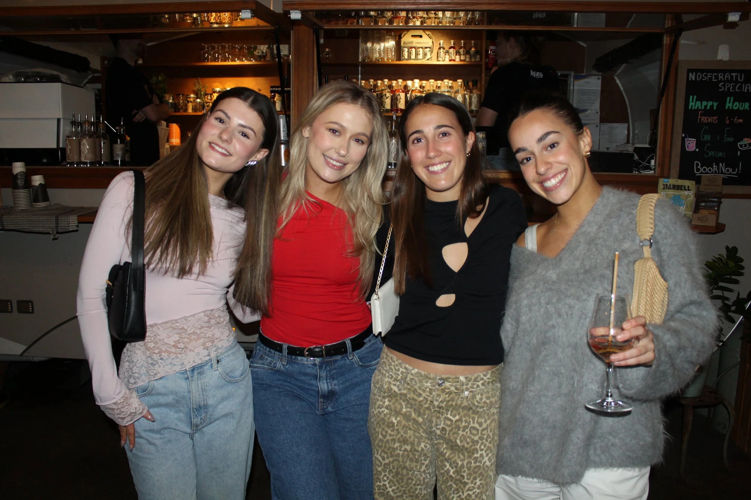 Four young women smiling and posing together in a bar or restaurant setting, with drinks in hand and a bar with bottles behind them.