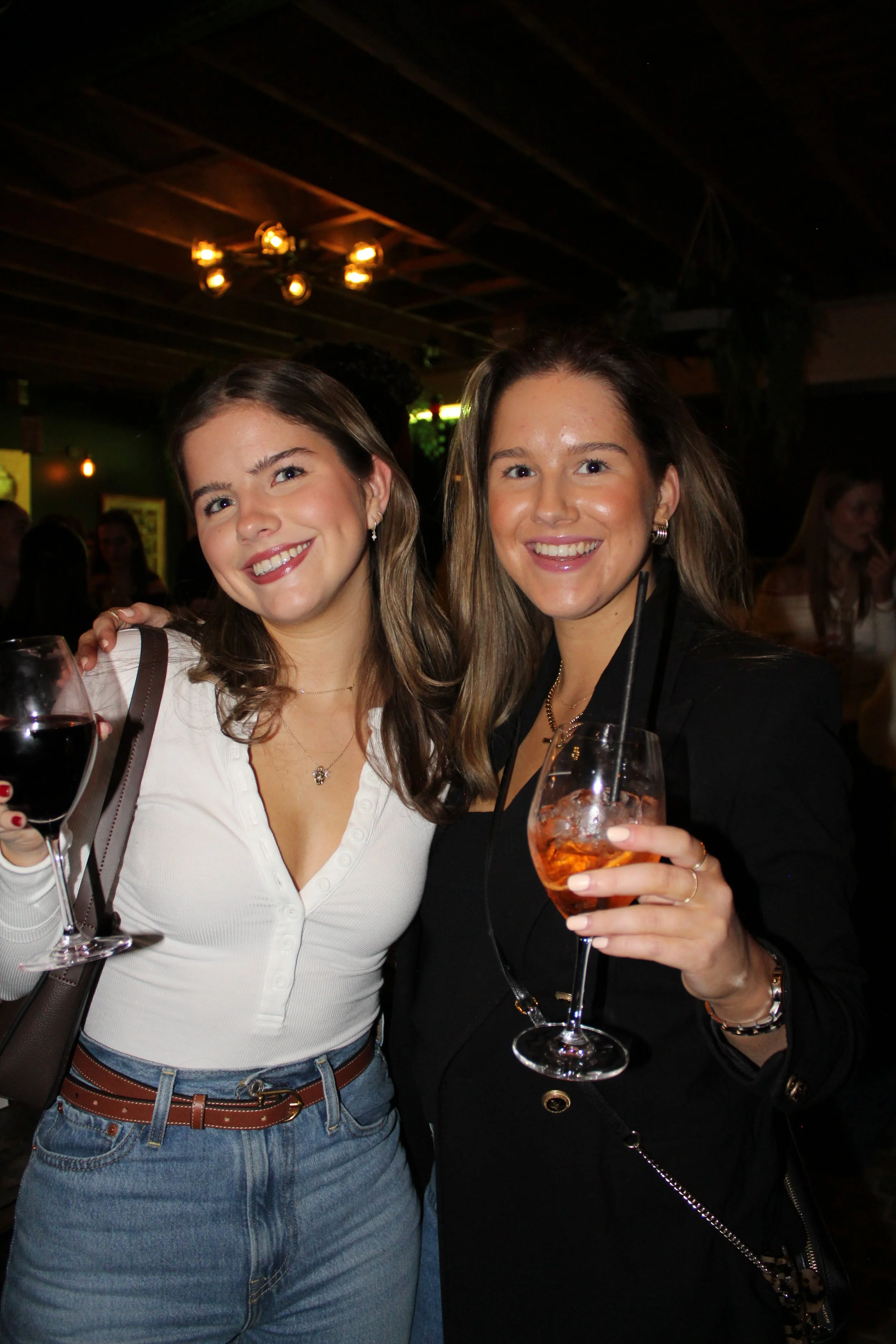 Two women smiling and holding drinks at a bar or restaurant, with one holding a glass of red wine and the other holding a cocktail.