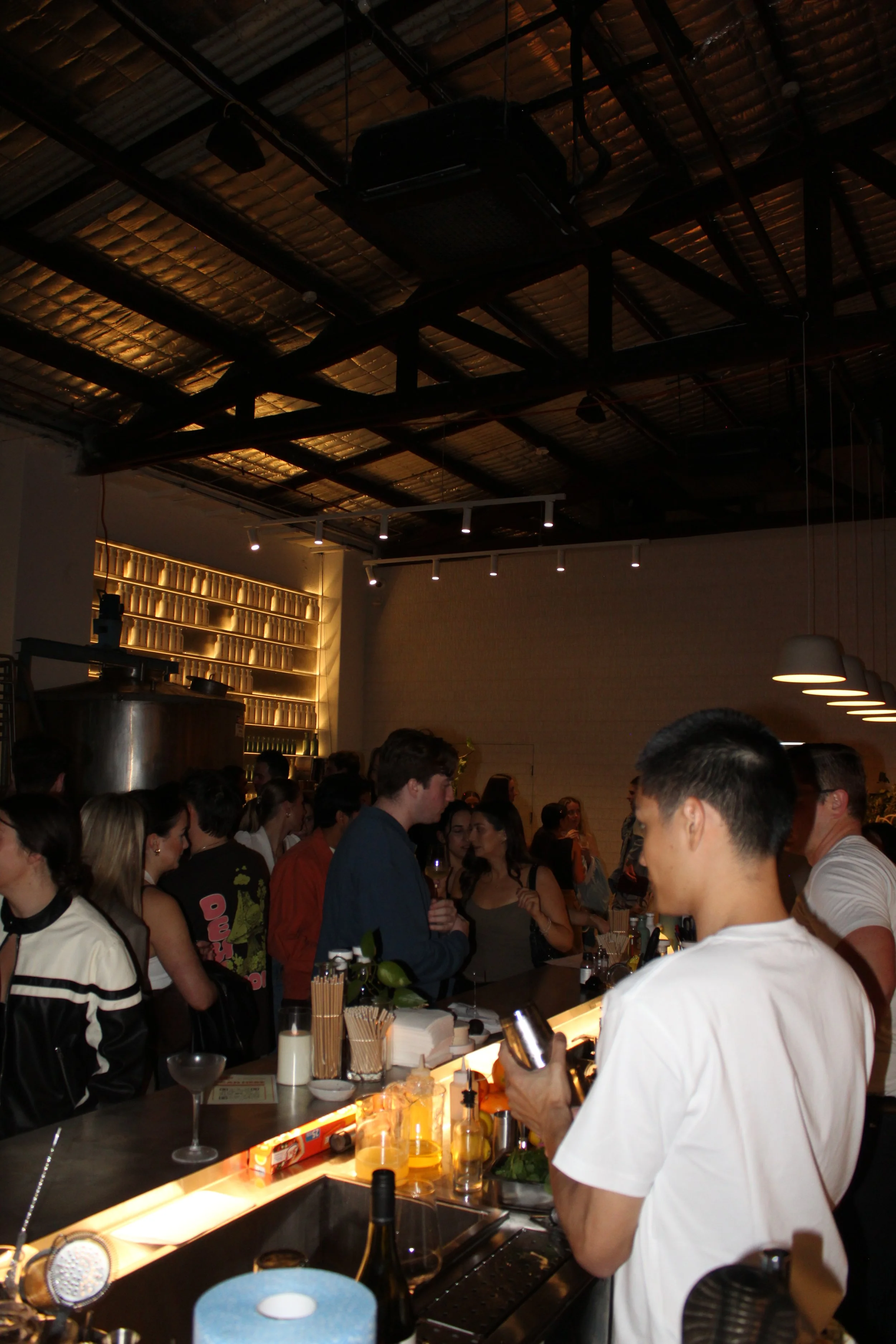 Crowded bar scene with people socializing and bartenders preparing drinks in a dimly lit setting with modern decor.