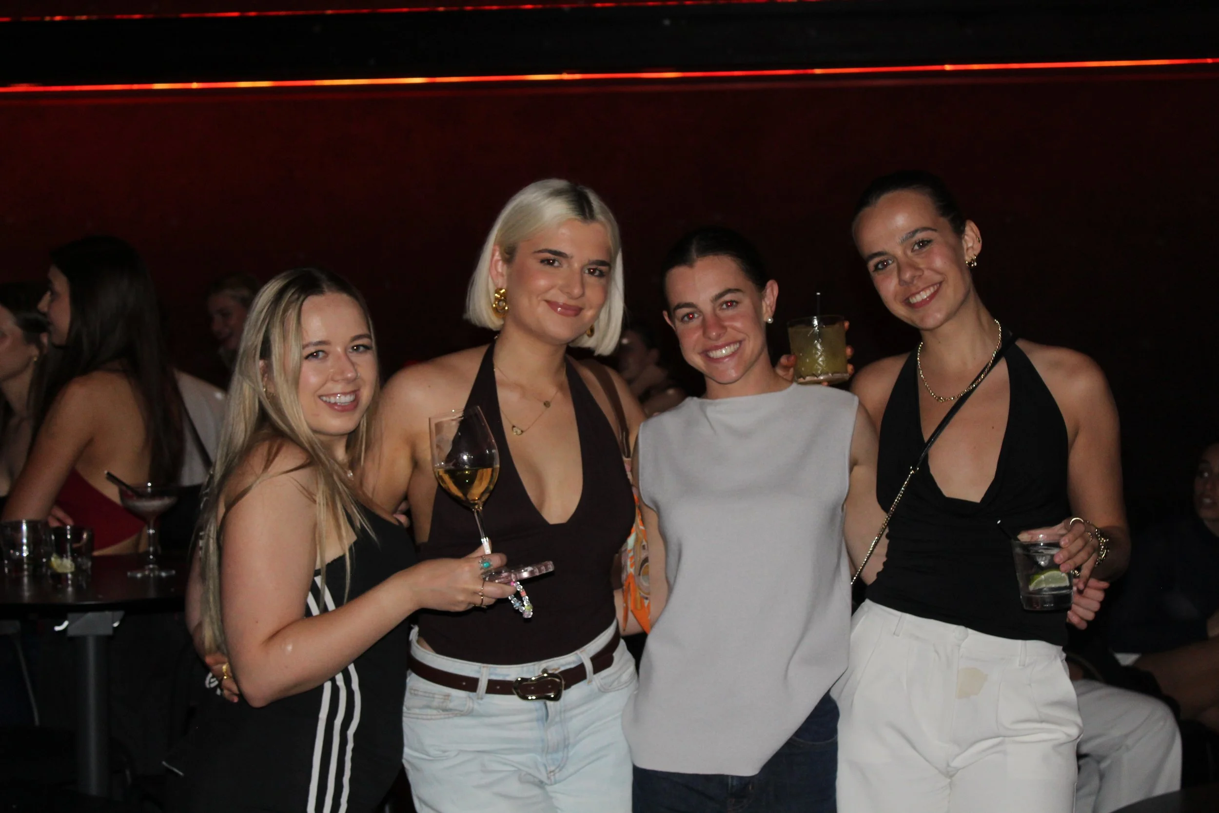 Four women standing together at a social event, holding drinks and smiling at the camera. They are in a dimly lit indoor setting with other people and drinks visible in the background.