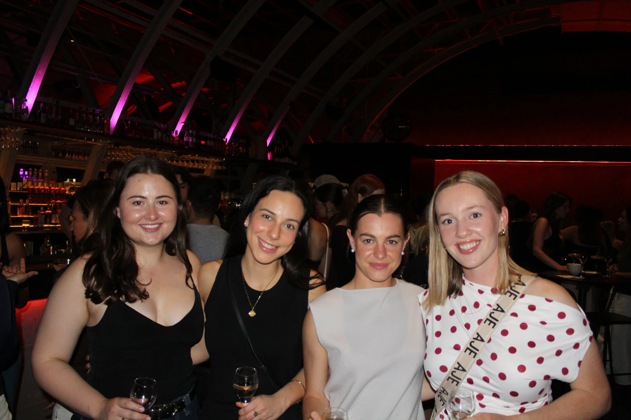 Four women standing closely together at a social event, smiling, with a bar and other people in the background.