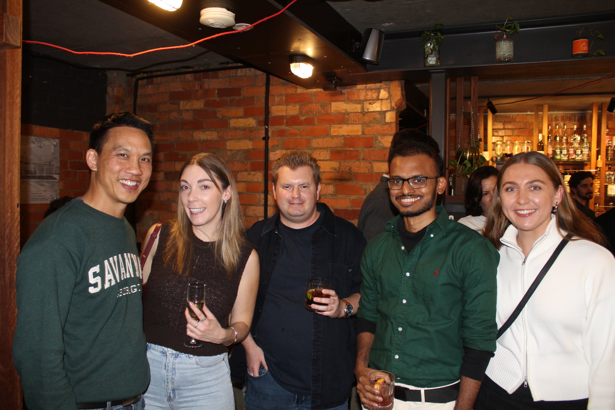 A group of six diverse people smiling at a bar or restaurant, holding drinks. They are standing close together in front of a brick wall and bar shelves with bottles, in a warm, casual setting.