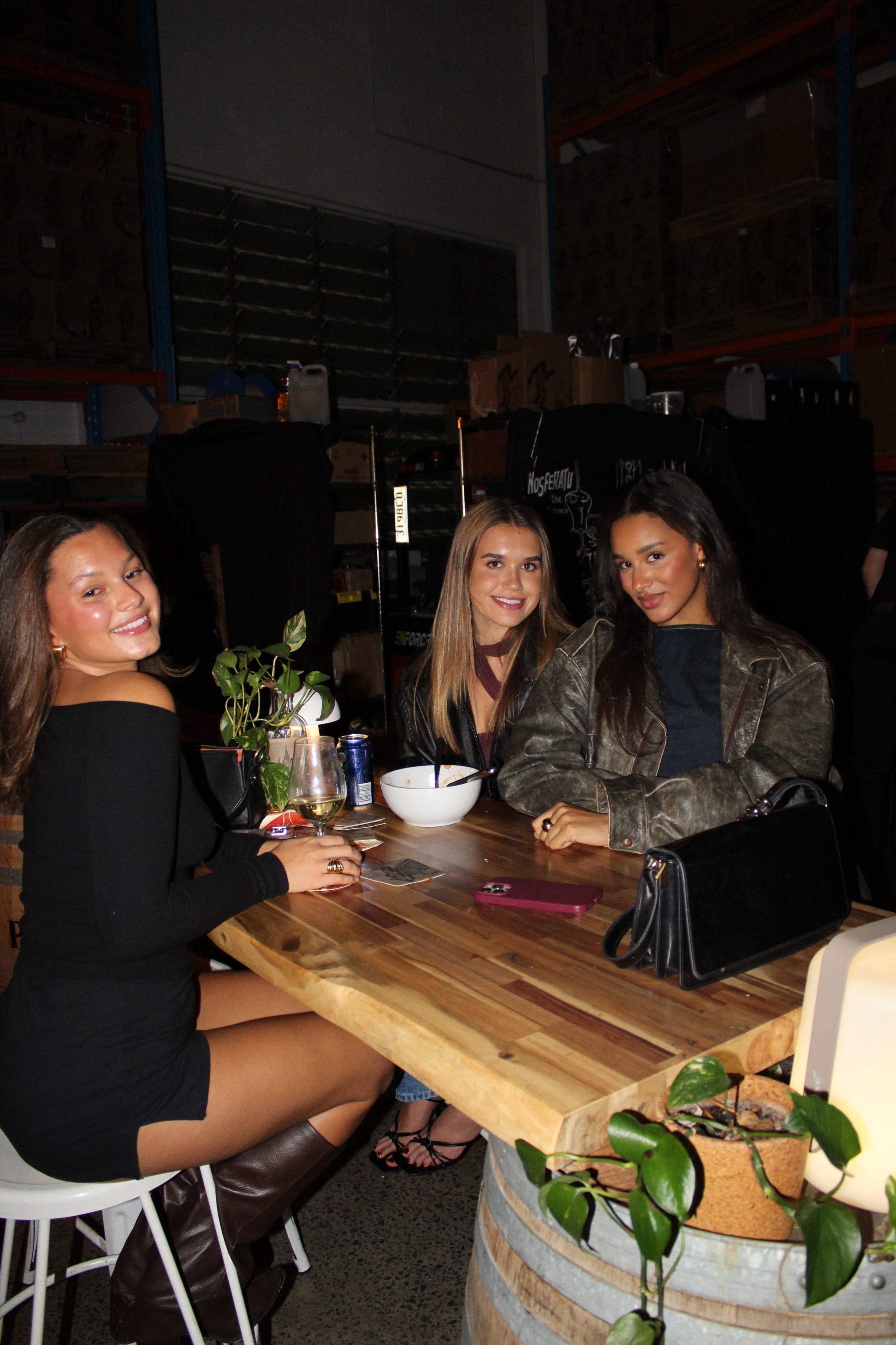 Three women sitting at a wooden table in a warehouse-style setting, smiling at the camera. One has long brown hair, another has shoulder-length light brown hair, and the third has long black hair. The table has a bowl, a glass of white wine, a potted