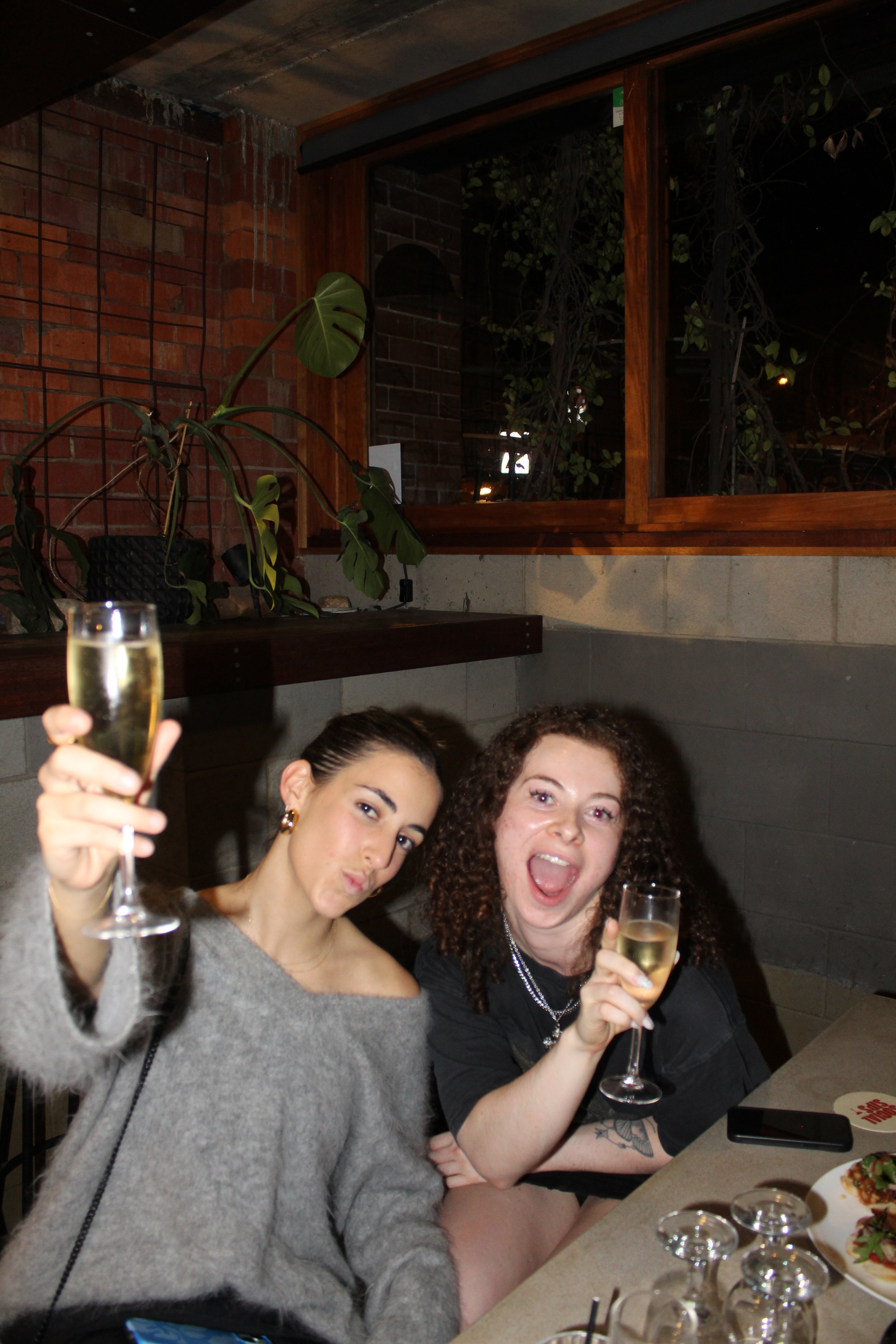 Two women raising glasses of champagne at a celebration in a restaurant or bar. One woman has dark hair and the other has curly red hair. They are sitting at a table with food and drinks, smiling and enjoying the moment.