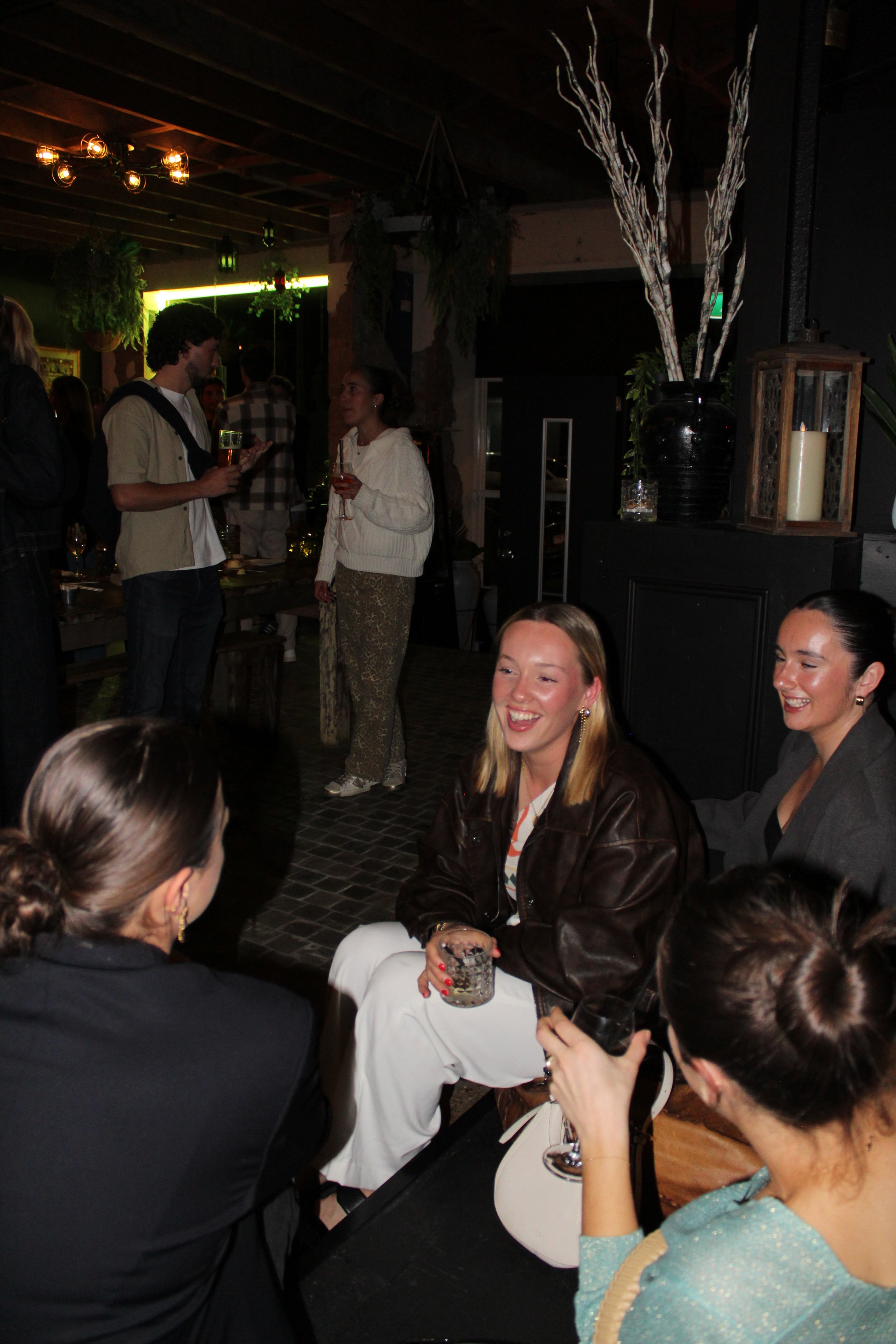 Group of women sitting and talking at a social gathering in a dimly lit indoor venue, with other people in the background holding drinks.
