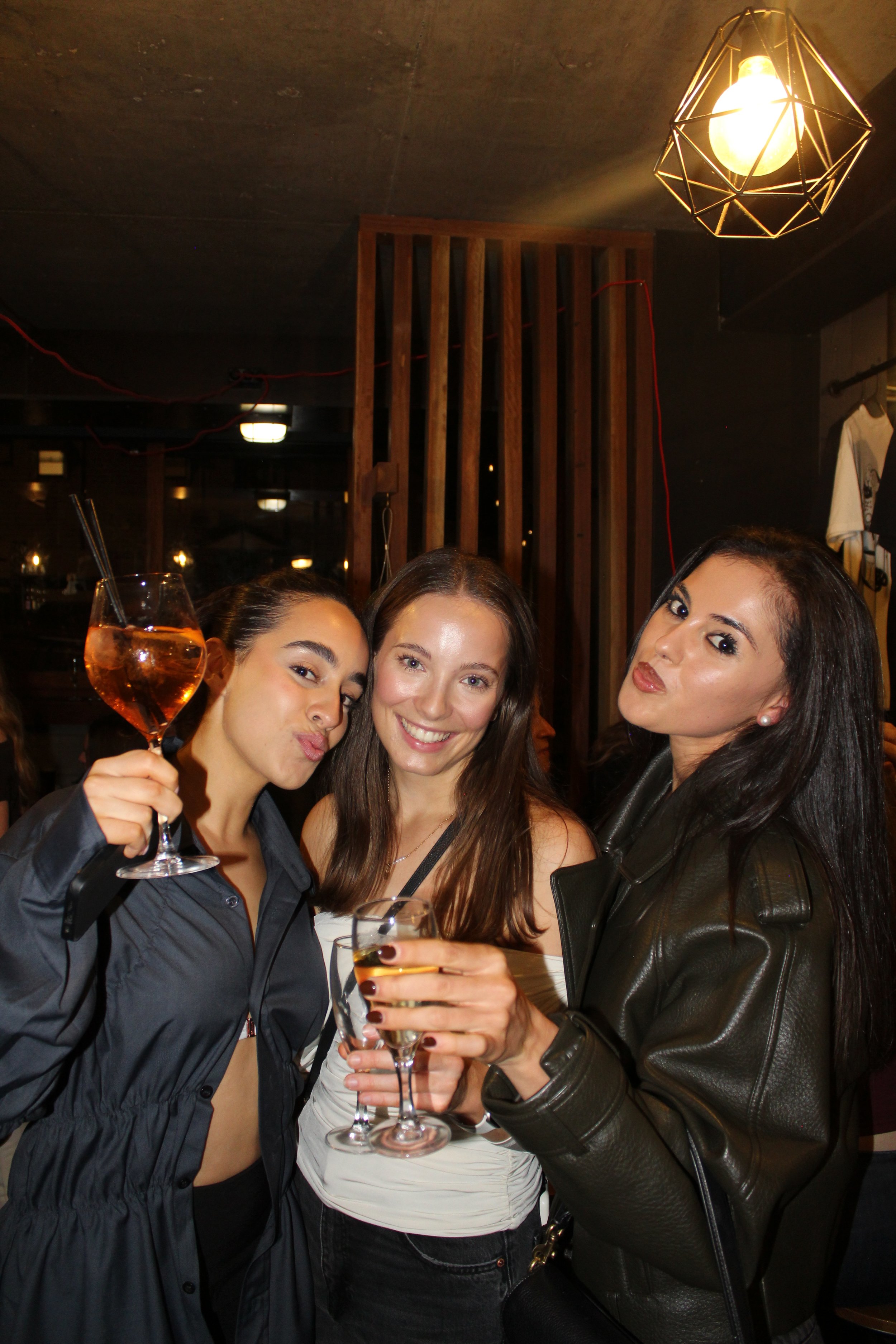 Three women smiling and holding drinks in a dimly lit bar or restaurant. One woman on the left is holding a glass of rosé wine, the woman in the middle has a white drink, and the woman on the right has a glass of champagne or sparkling wine. They are