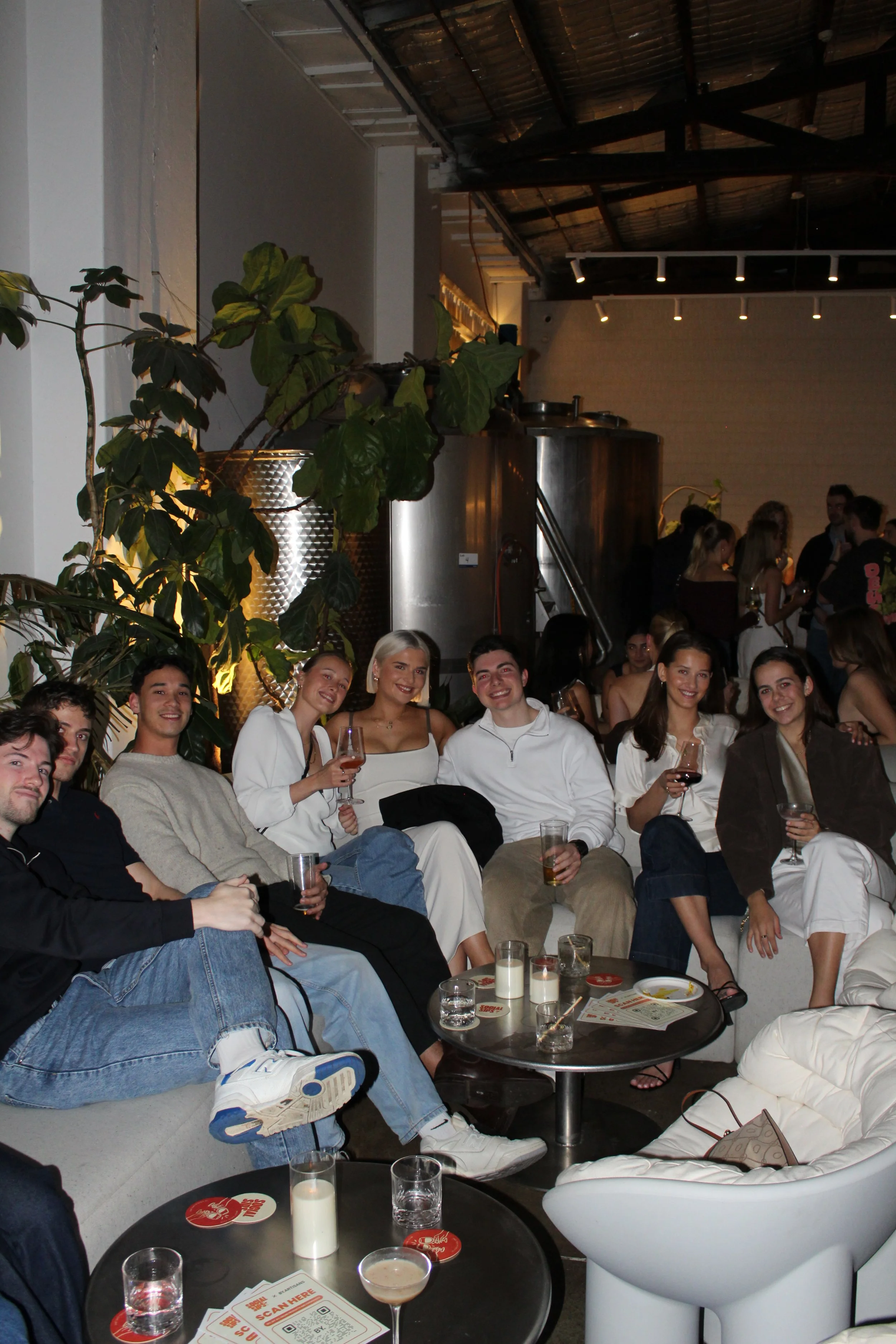 Group of people sitting on couches at a social gathering in a dimly lit indoor venue, holding drinks and smiling.