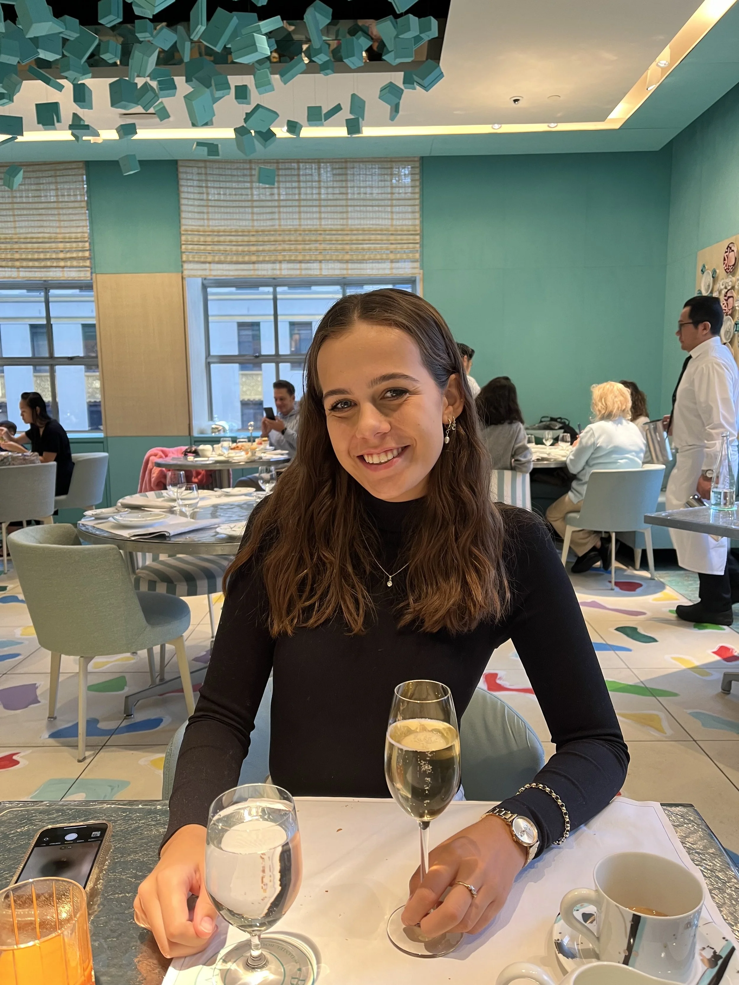 A man with dark, curly hair, a beard, and clear glasses, wearing a dark green sweater, sitting indoors with a neutral expression.