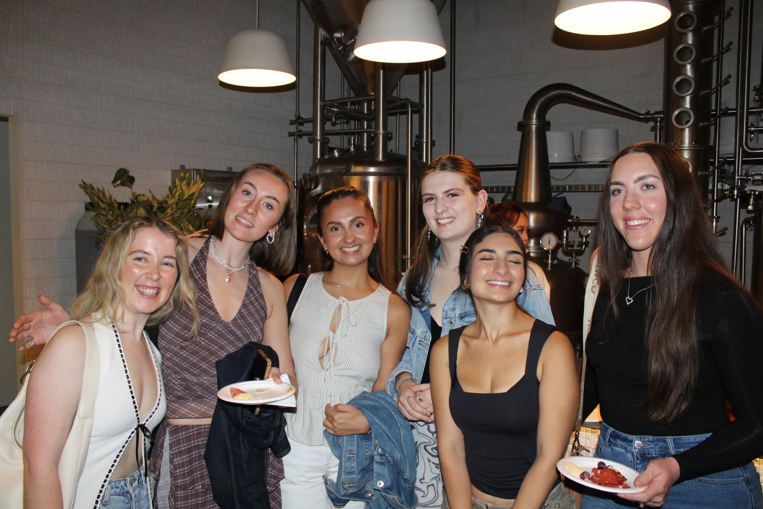 Group of smiling young women at a social gathering, holding plates with food, indoors with industrial-style equipment in the background.