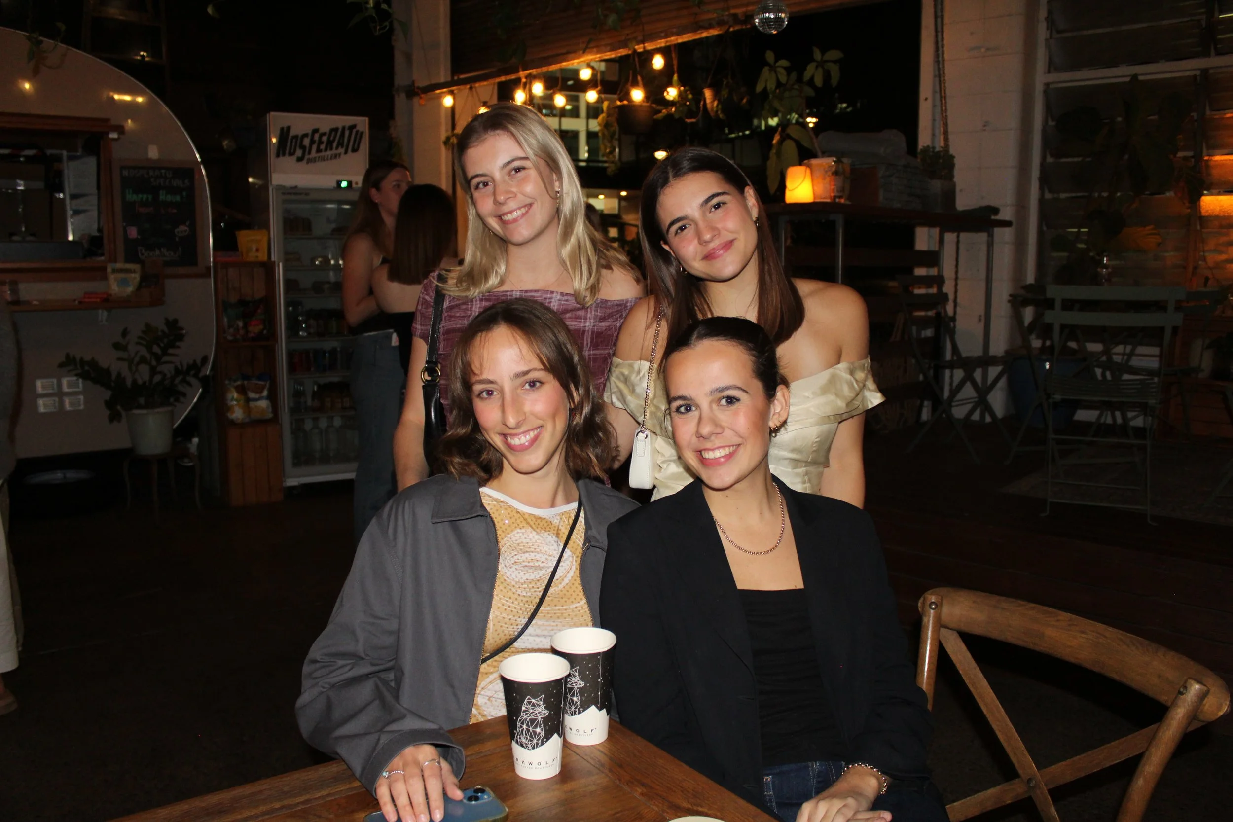 Group of five young women smiling at a social gathering at a restaurant or bar. Two women are seated at a table with two paper cups, and three women are standing behind them. The setting has warm lighting and a cozy, modern decor.