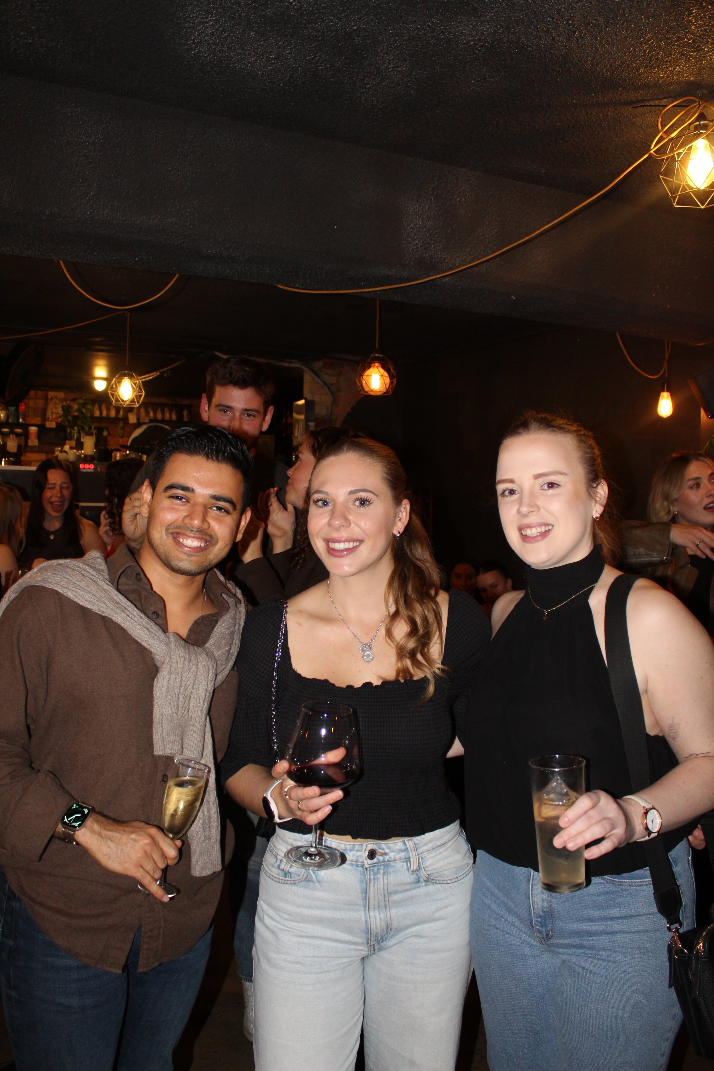 Group of four people smiling, holding drinks, inside a dimly lit bar with hanging pendant lights.