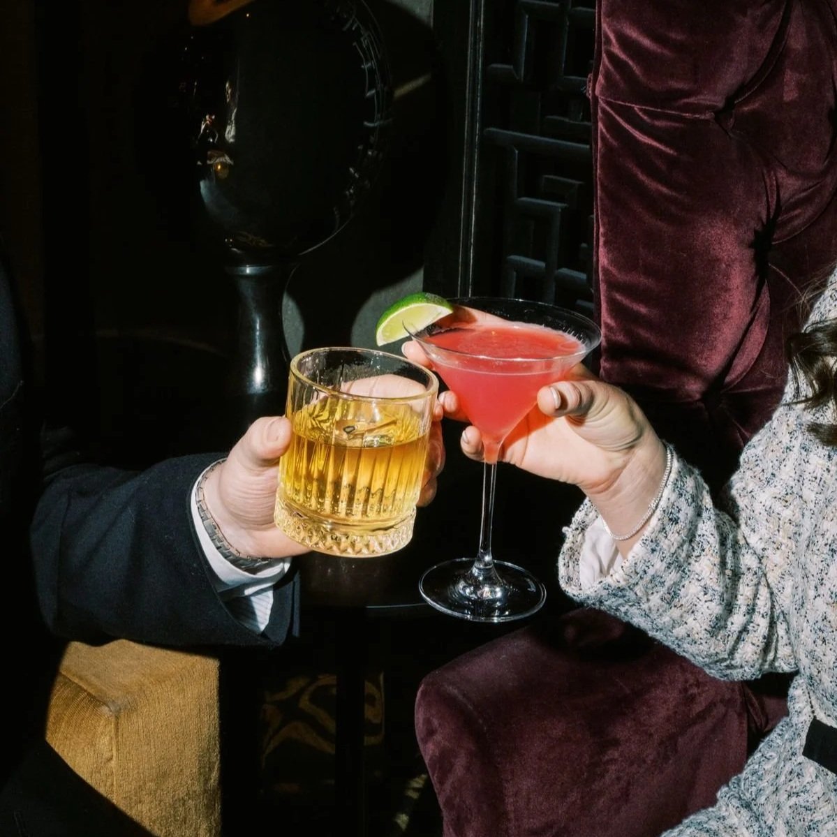 Two people holding glasses of drinks toasting at a bar, one with a cocktail garnished with lime and the other with a glass of whiskey.