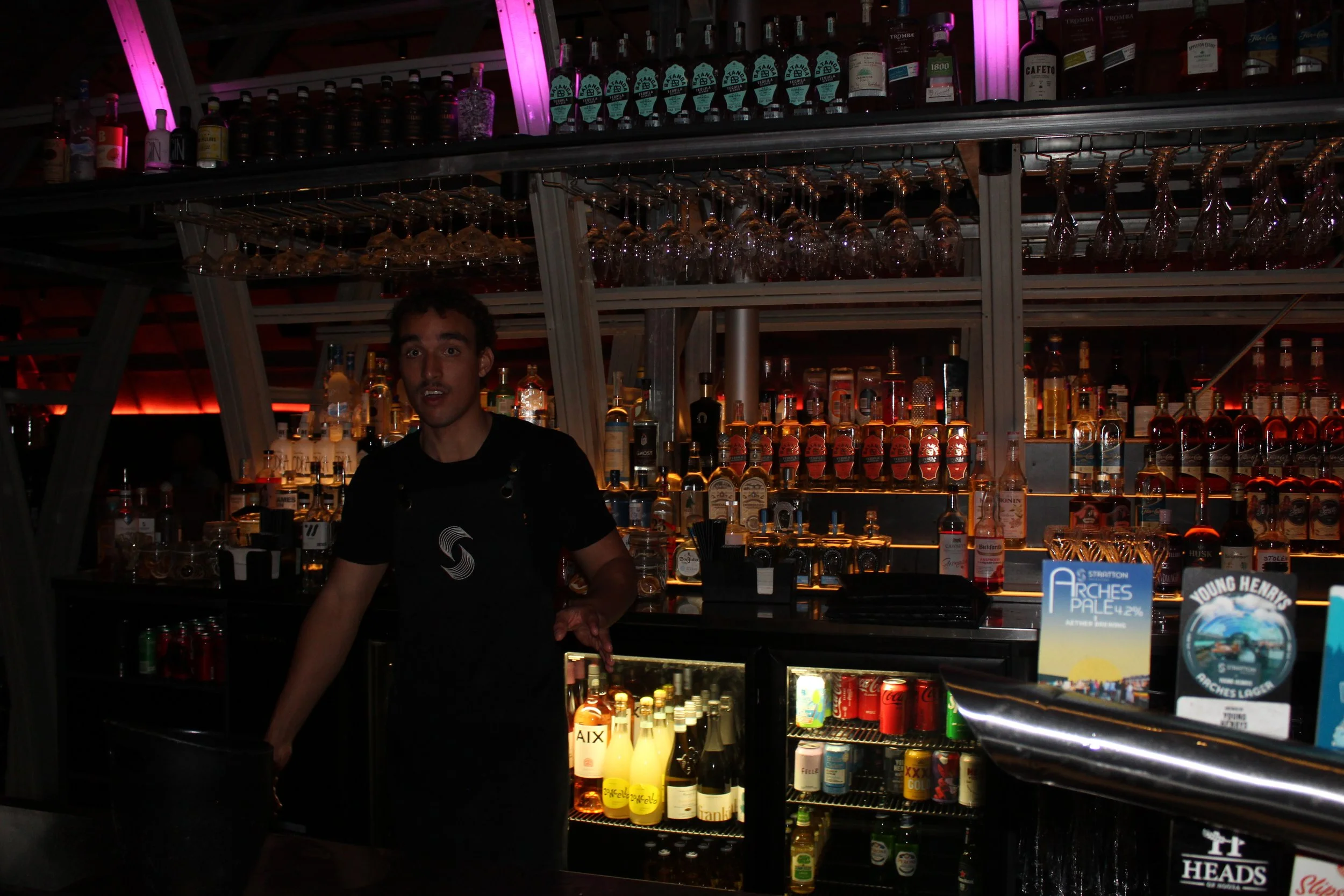 A bartender behind a bar with shelves of liquor bottles and hanging wine glasses, under purple and red lighting.