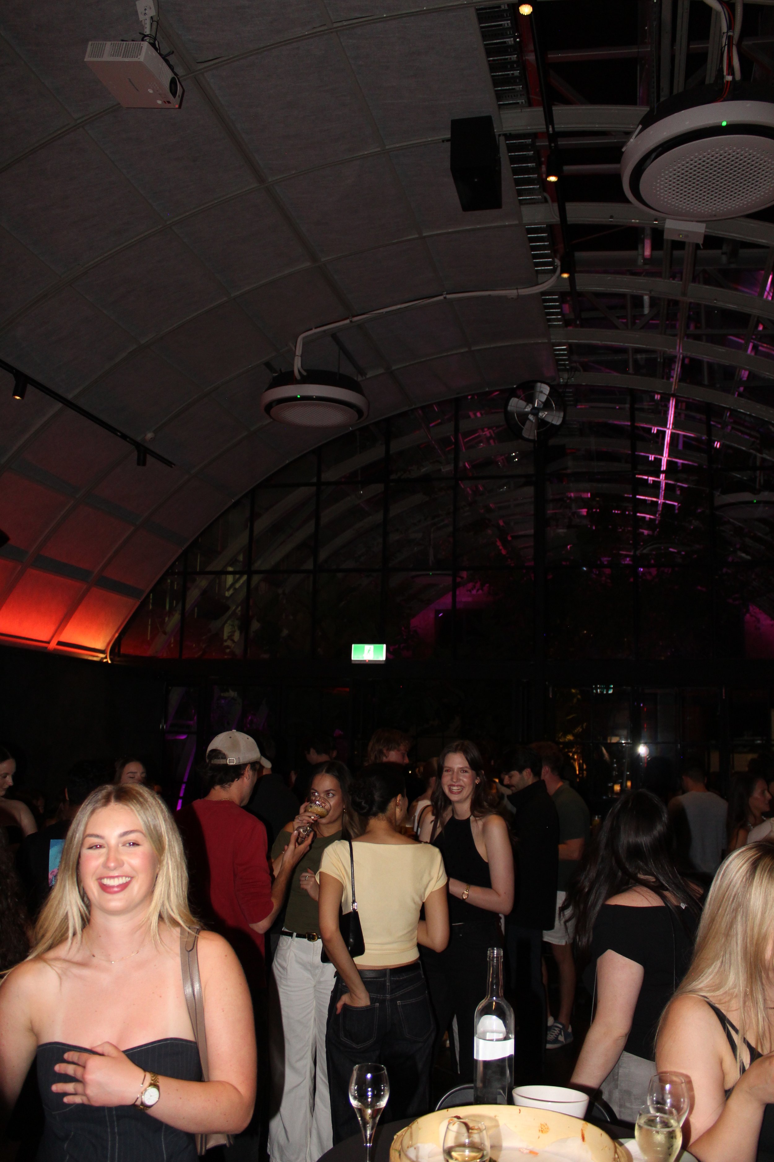 People socializing at a party or nightclub with drinks, smiling and talking under a large, arched ceiling with colorful lighting.