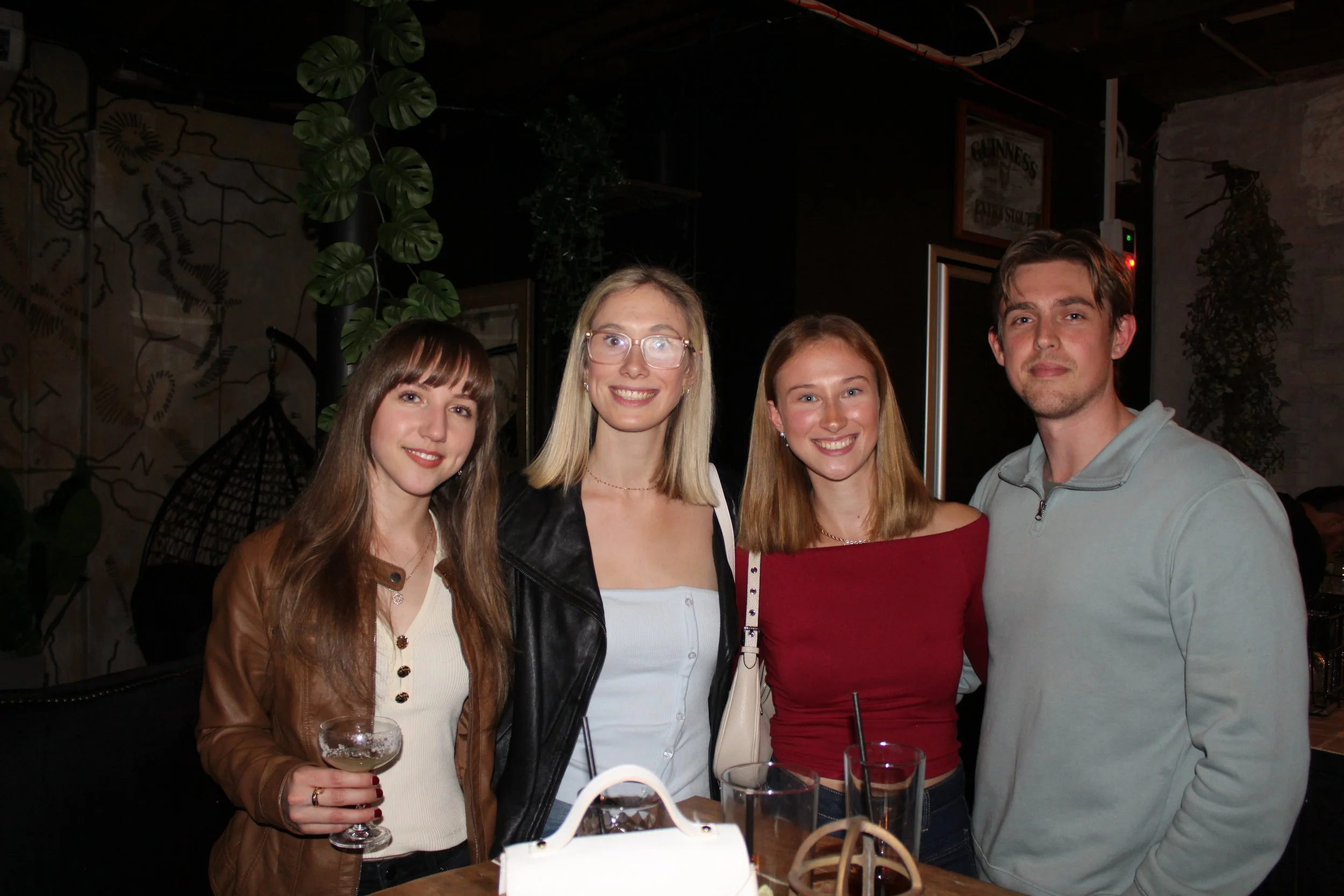 Four friends smiling and posing for a photo at a bar or restaurant, with drinks in hand, in a dimly lit indoor setting with plants and artwork in the background.