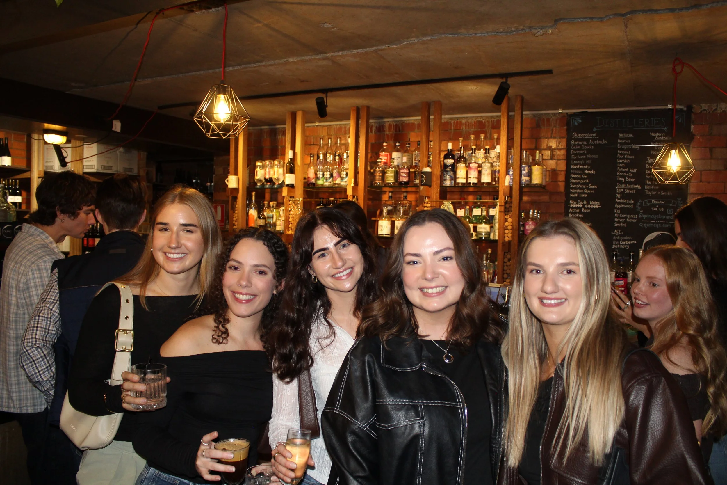 Group of young women smiling and holding drinks at a bar or social gathering, with a background of liquor bottles and a chalkboard menu.