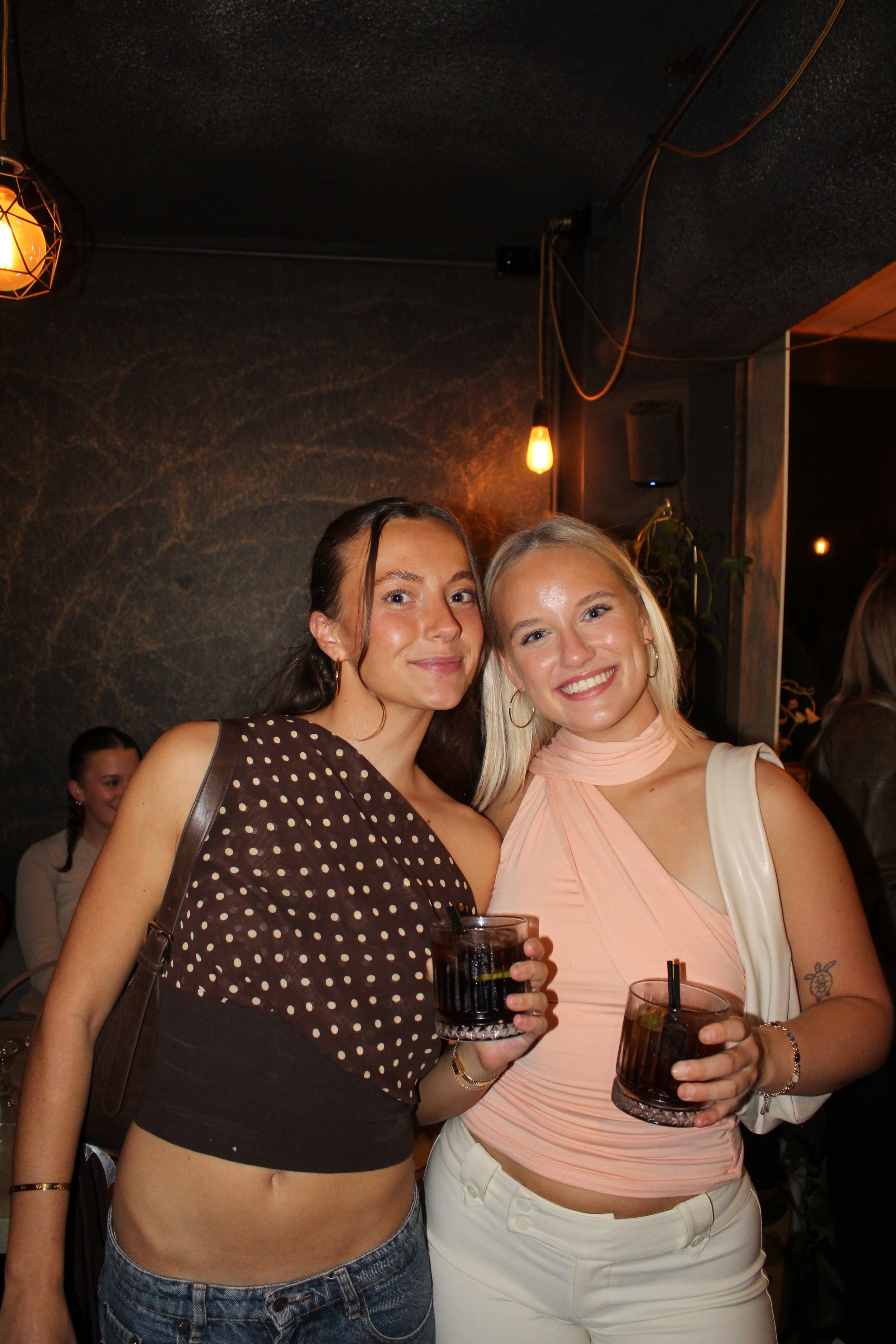 Two women are smiling and posing for a photo in a dimly lit bar or restaurant, holding glasses of dark-colored drinks with ice. The woman on the left has long dark hair, is wearing a brown polka-dot top, and has a small purse. The woman on the right 
