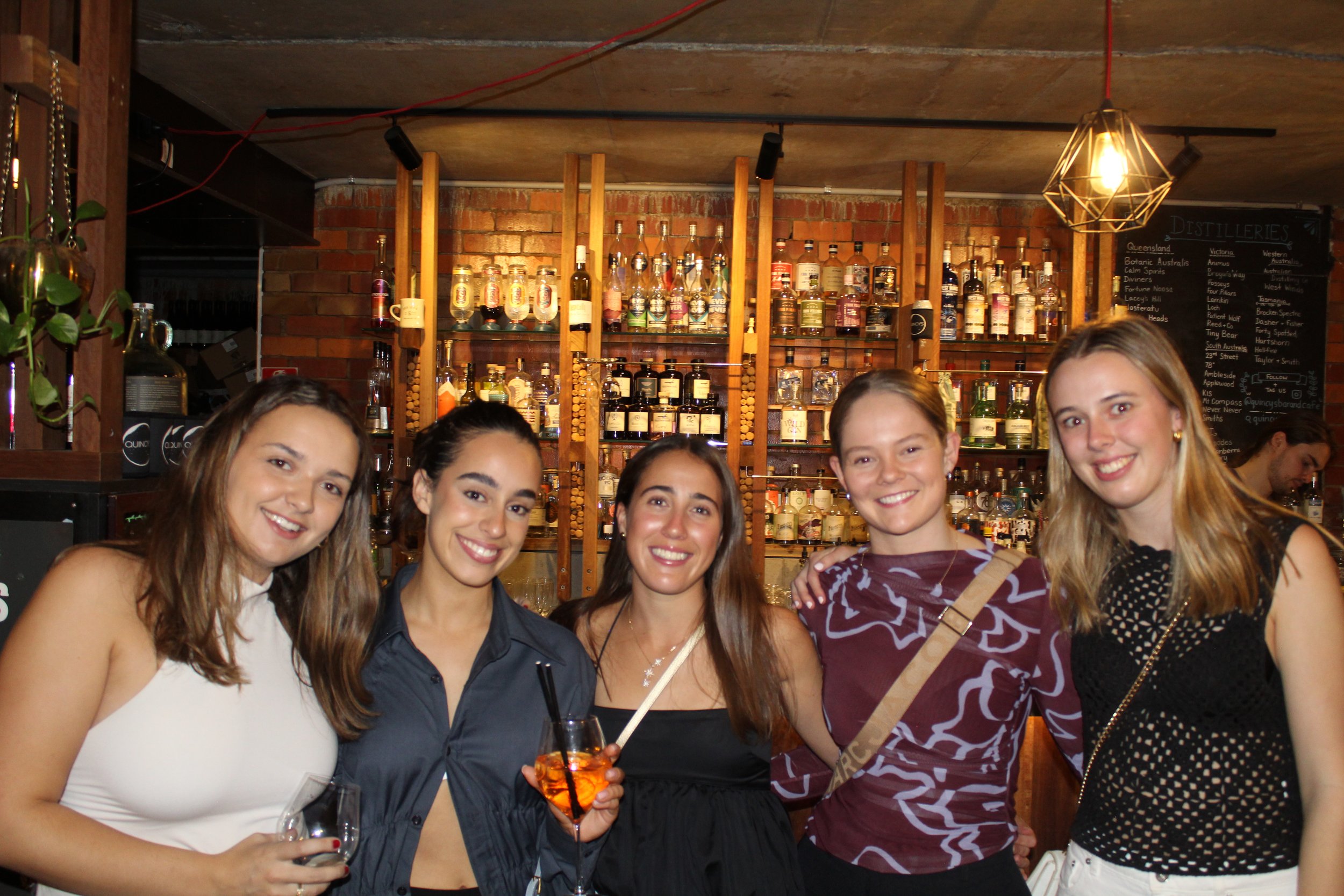 Group of young women smiling and enjoying drinks at a bar with a wooden shelving back displaying various bottles of alcohol.