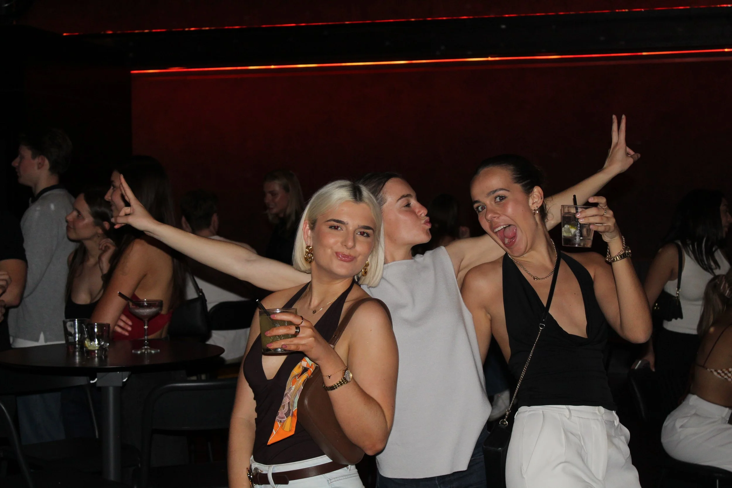 Three women having fun at a bar or club, holding cocktails, with other people in the background. The woman in the middle is making a kissy face, the woman on the right is smiling and raising her hand, and the woman on the left is smiling with a drink