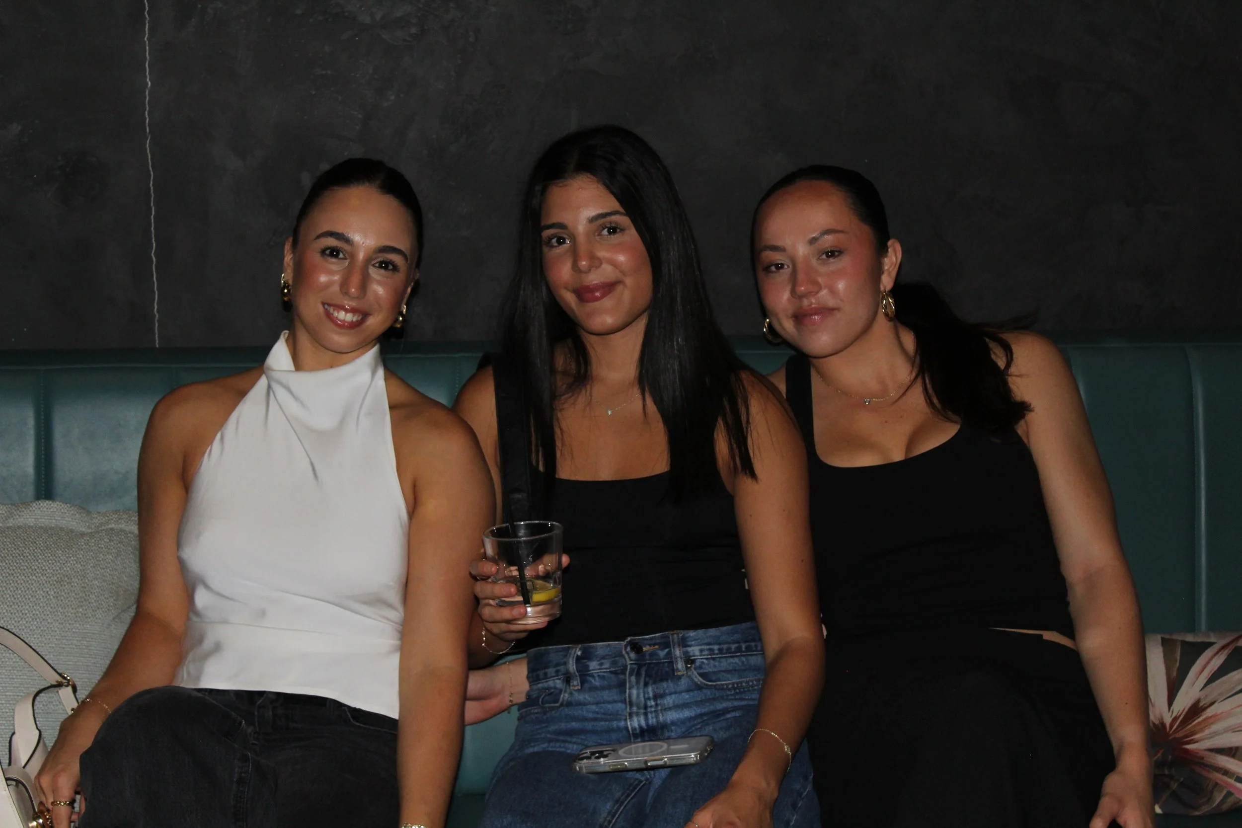 Three women sitting together on a teal upholstered bench in a dark indoor setting, smiling at the camera. The woman on the left is wearing a white sleeveless top and black pants, the woman in the middle is wearing a black top and jeans and holding a 